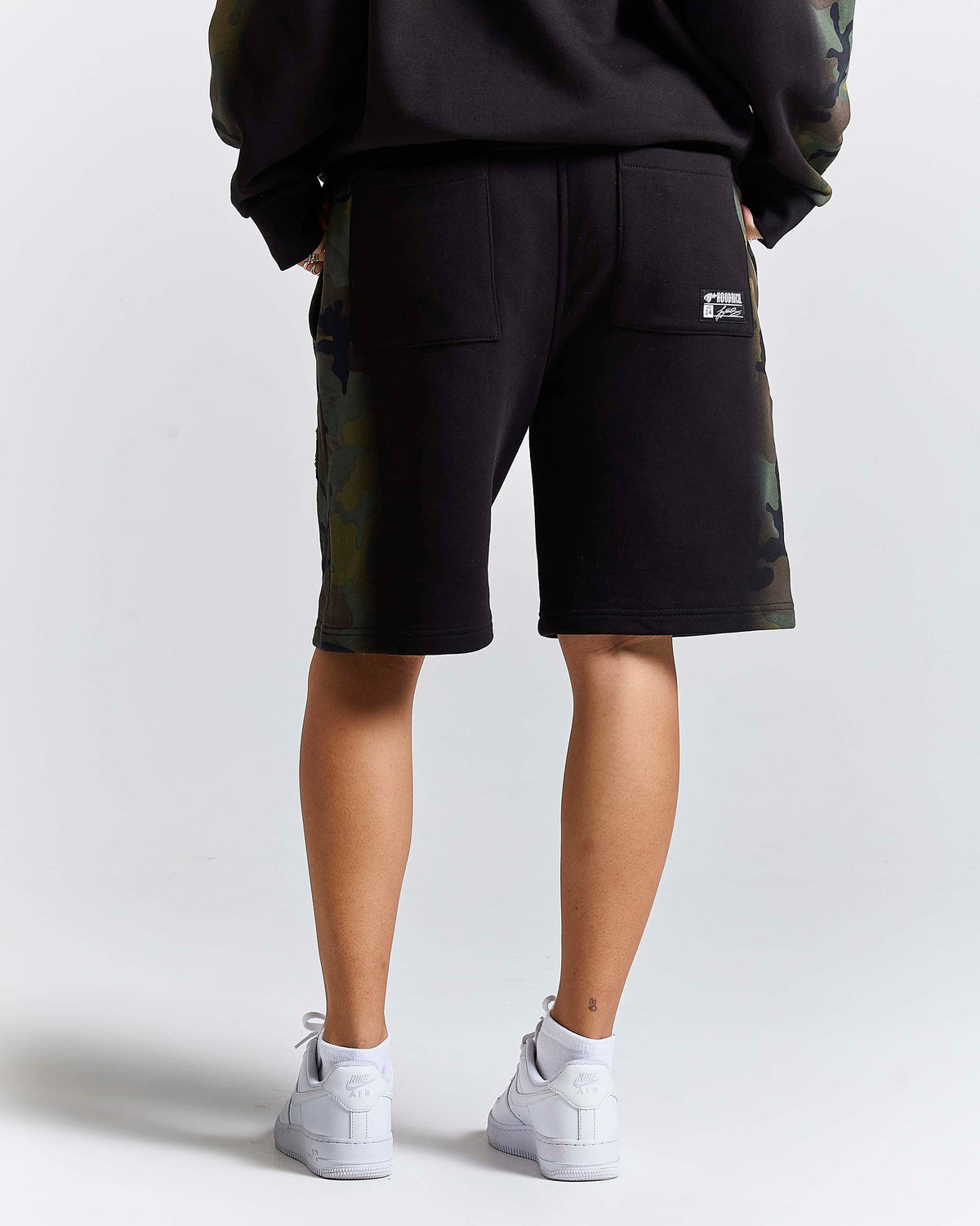 A person shown from the back wearing black shorts with camouflage side panels, a black sweatshirt, and white sneakers against a light gray studio background.
