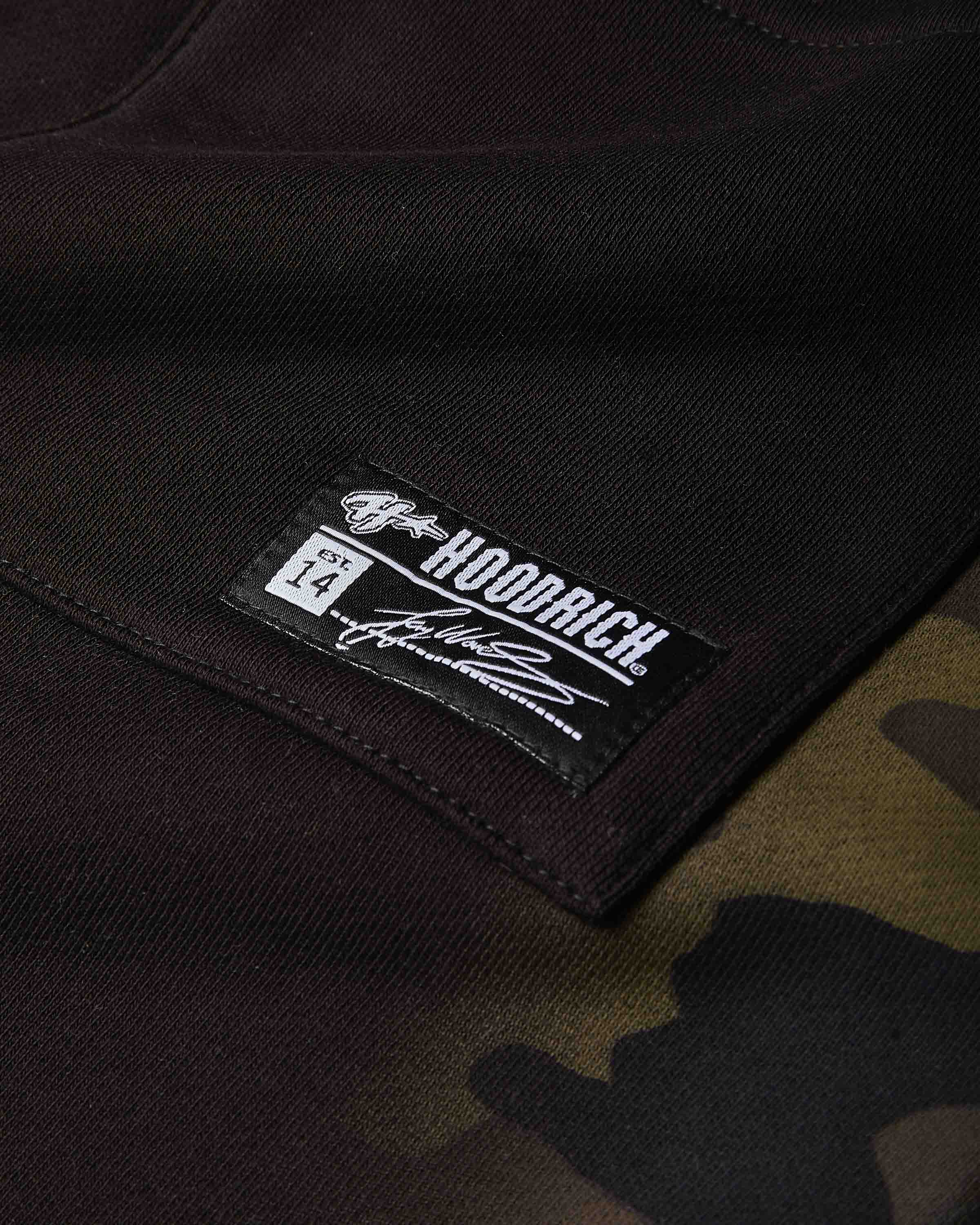Close-up of a black HOODRICH brand patch sewn onto a dark brown and black camouflage hoodie.