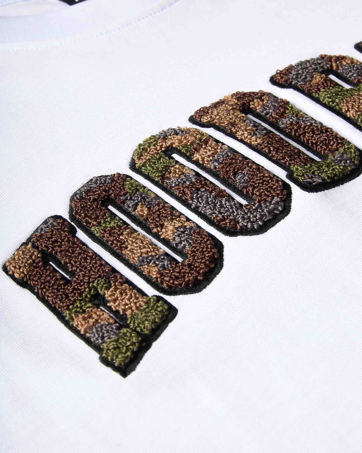 Close-up of a white t-shirt featuring a large, textured embroidered graphic of 'HOODRICH' in a camouflage pattern with black outlines.