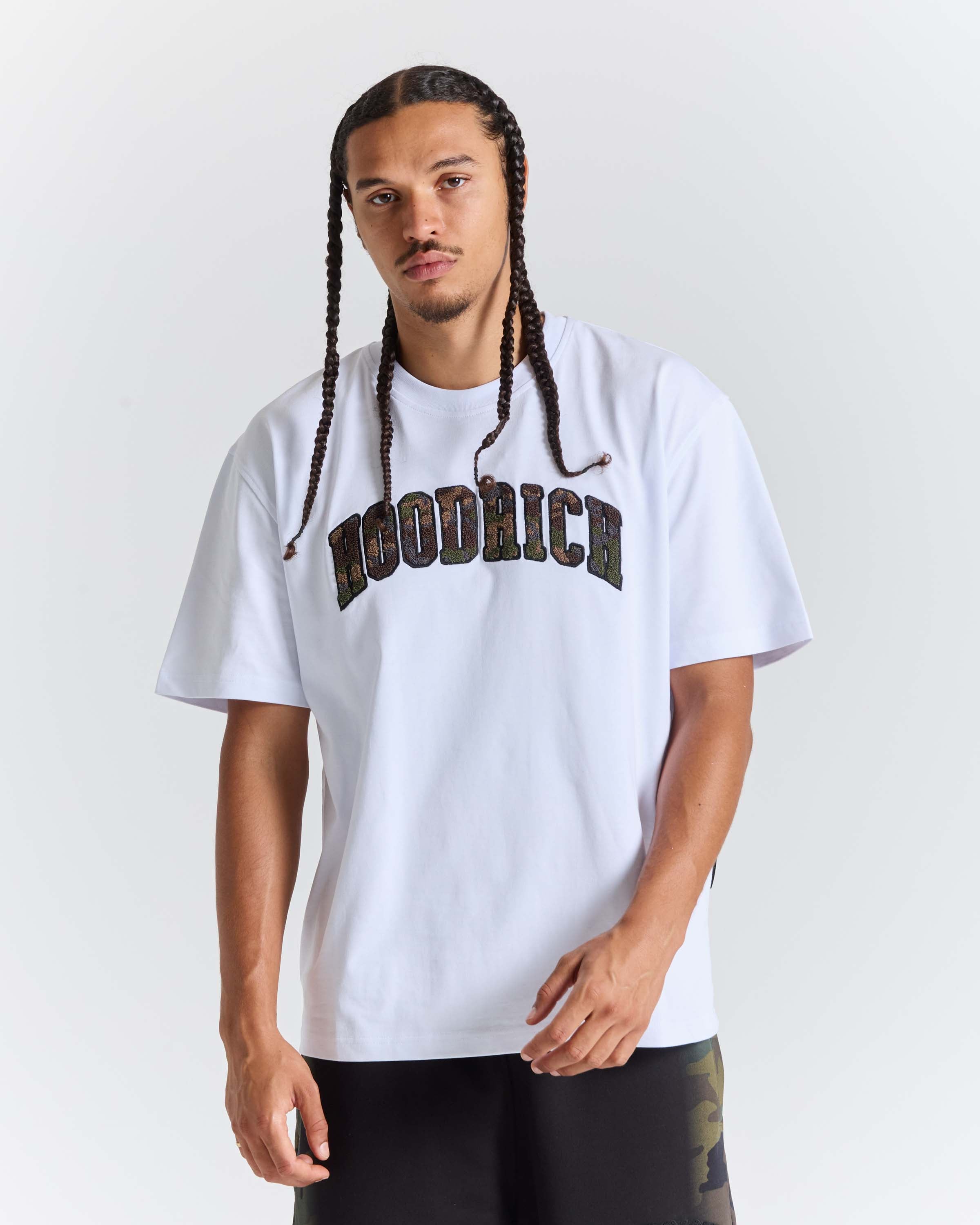 A man with long braids wearing a white Woodrich t-shirt with a dark camouflage-style arched Hoodrich logo across the chest, paired with black pants, against a light gray studio background.