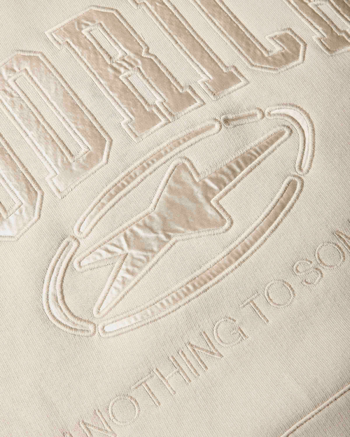 Close-up of a beige embroidered fabric with a large stylized letters and a logo outlined in raised stitching.