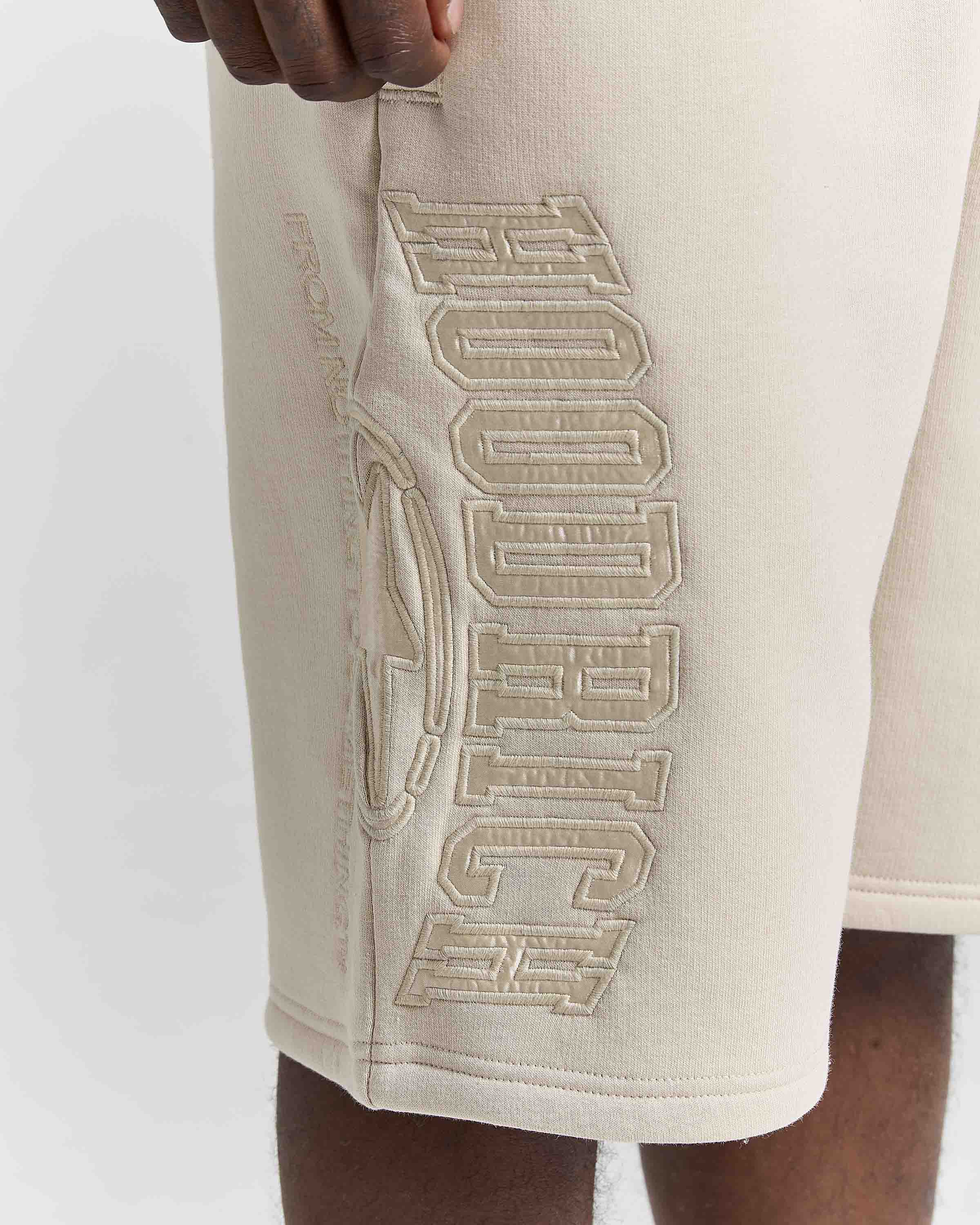 Close-up of a person wearing cream-colored athletic shorts with large tonal embroidered HOODRICH lettering on the side.