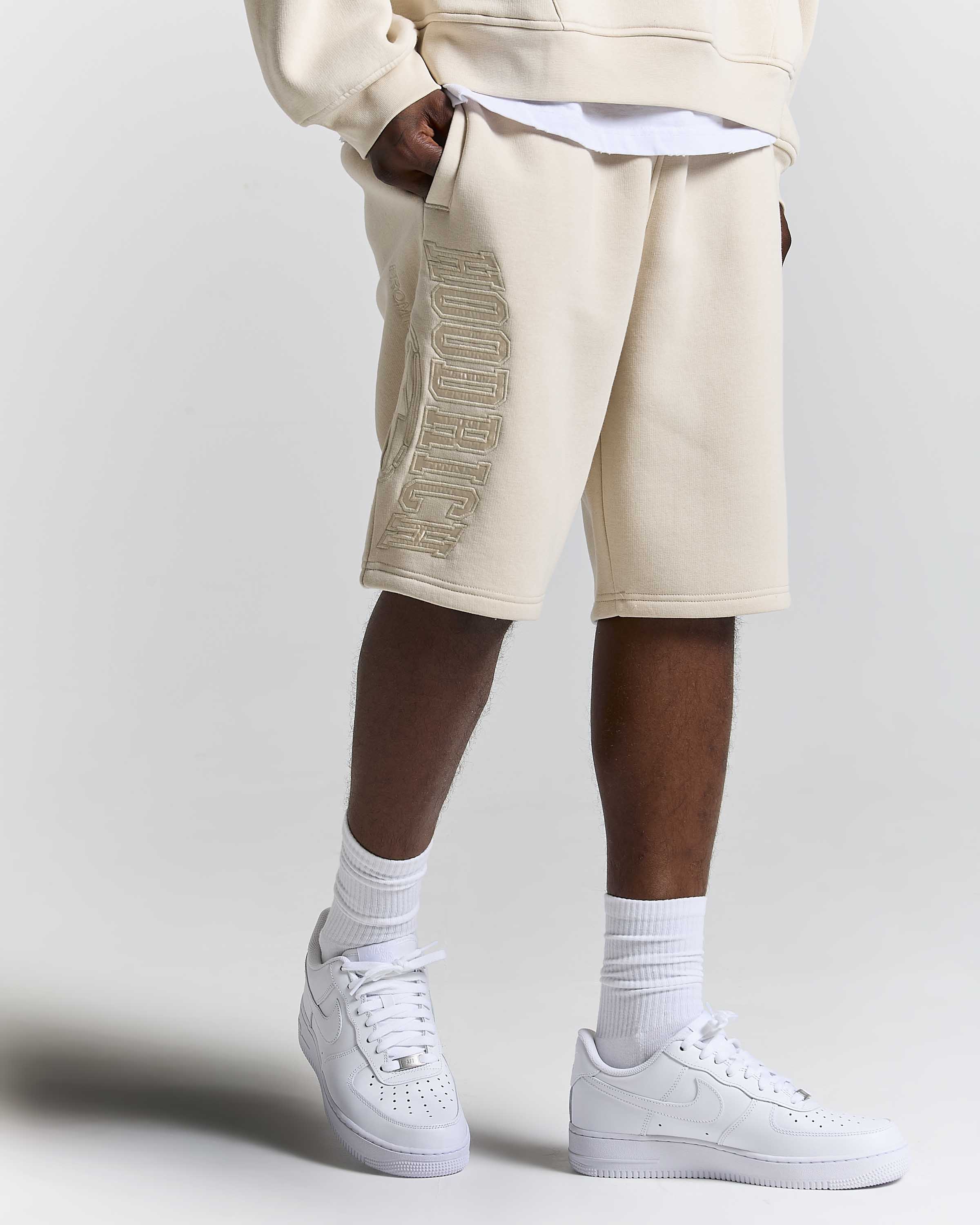 A person wearing beige sweat shorts with a metallic gold embroidered HOODRICH logo down the side, styled with a white t-shirt, white crew socks, and white Nike Air Force 1 sneakers against a light gray studio background.