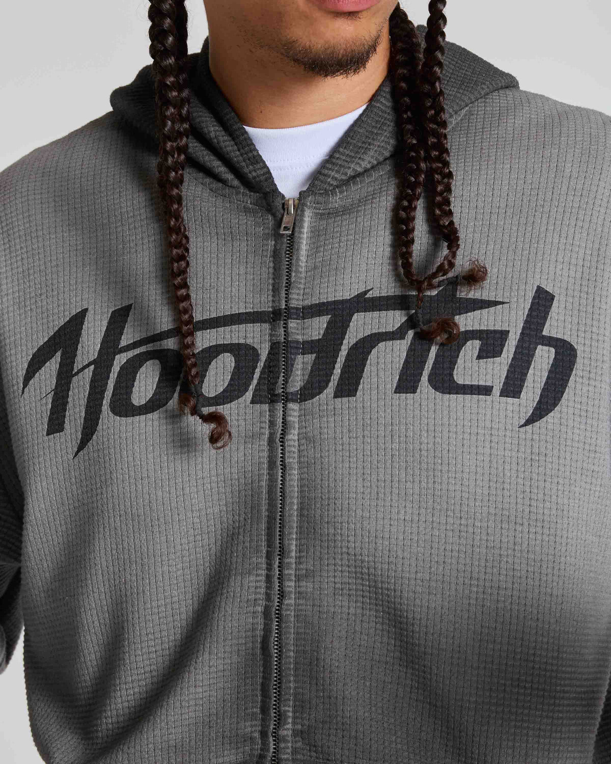 A close-up of a person wearing a gray waffle-knit zip-up hoodie with a large black 'Hoodrich' logo across the chest, over a white t-shirt. The person has dark brown braided hair hanging down the front.