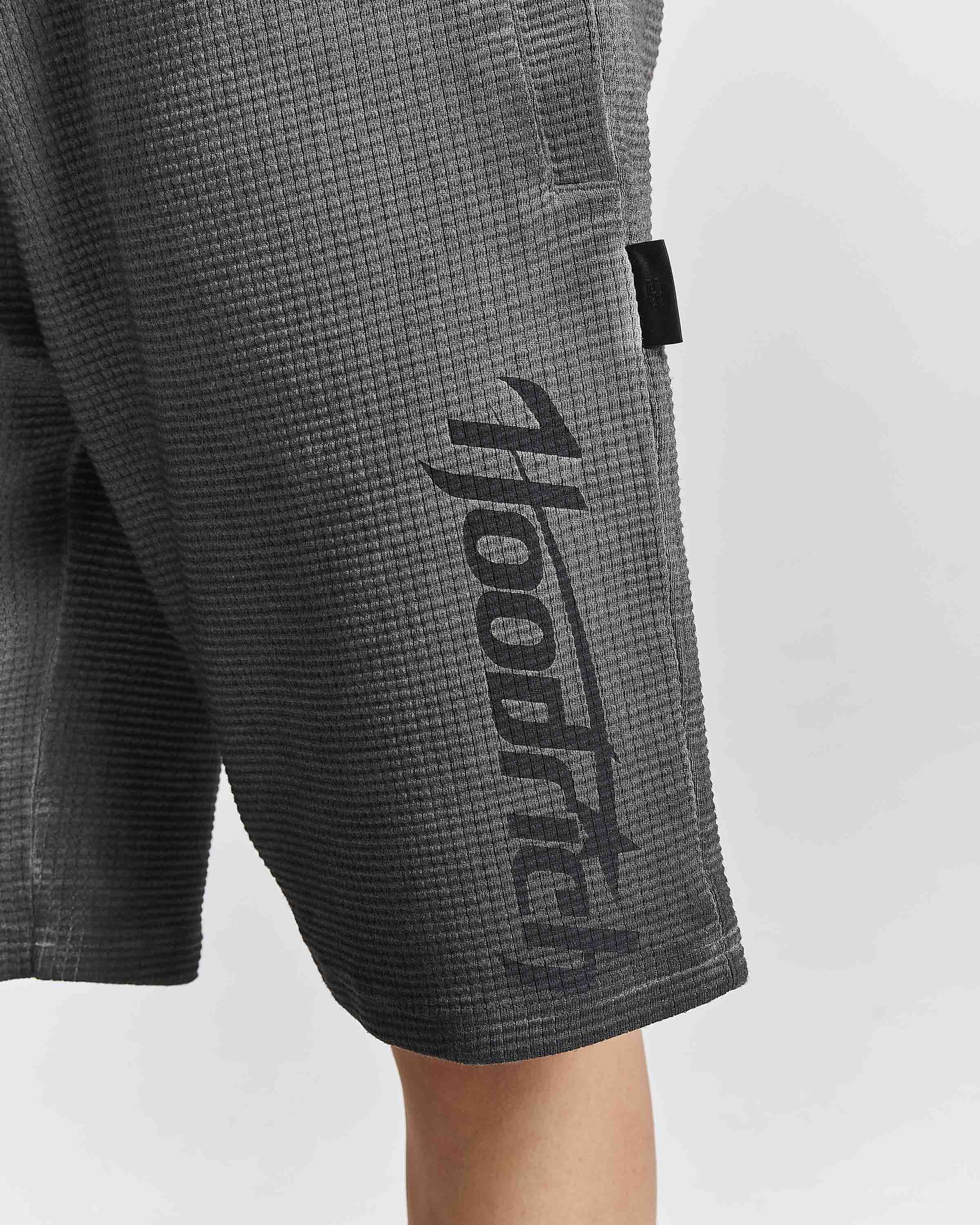 Close-up of dark gray waffle-knit shorts with a large black vertical 'hooded' logo printed on the side, worn by a model against a light gray background.