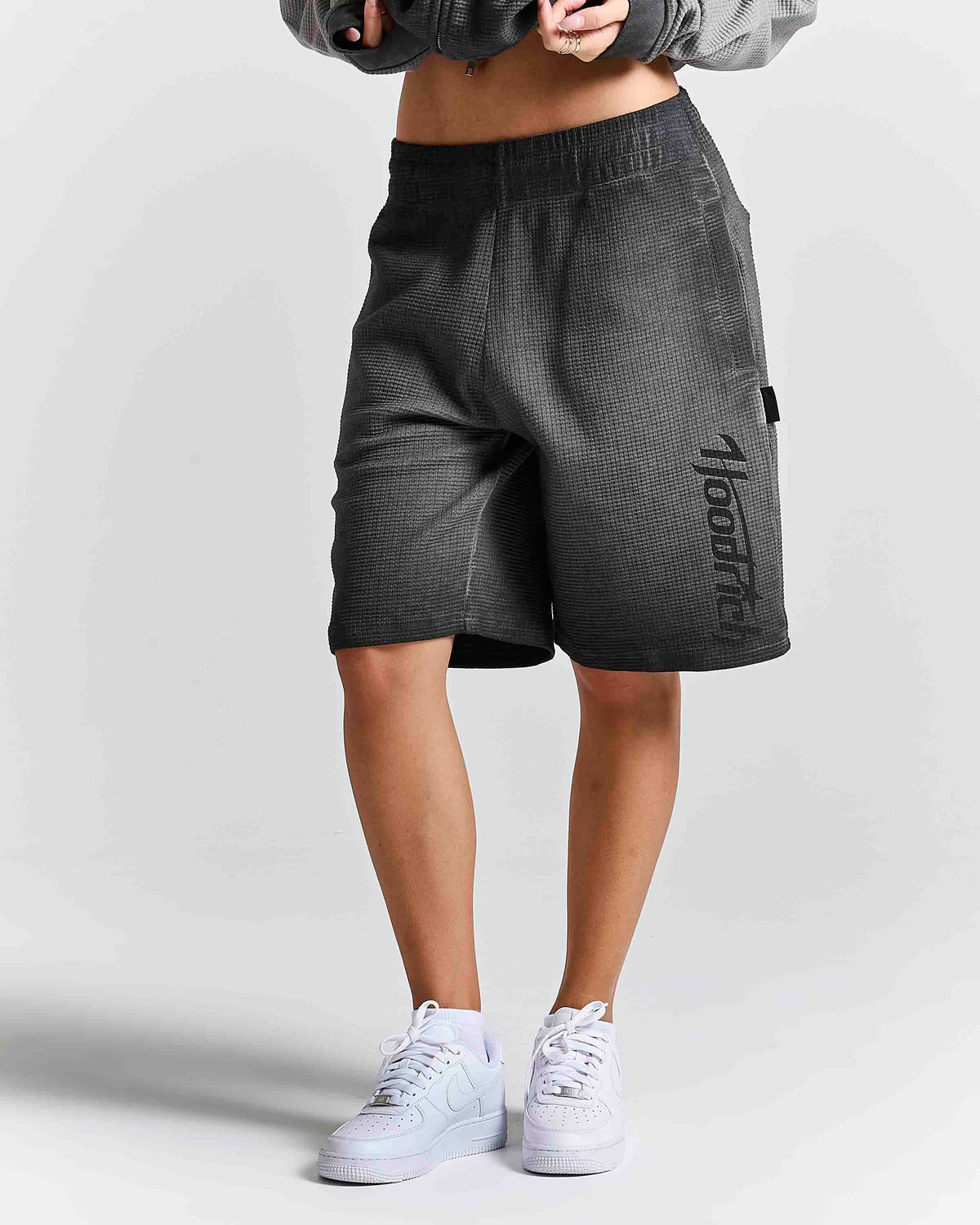 A person wearing dark gray textured athletic shorts with a vertical black 'Mood' logo on the side, paired with white sneakers and a gray cropped jacket, shown from the waist down against a light gray studio background.