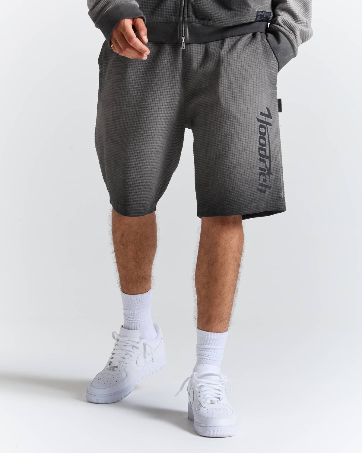 A person wearing dark gray textured athletic shorts with a vertical black 'hoodrich' logo on the left leg, styled with white crew socks and white sneakers against a light gray studio background.