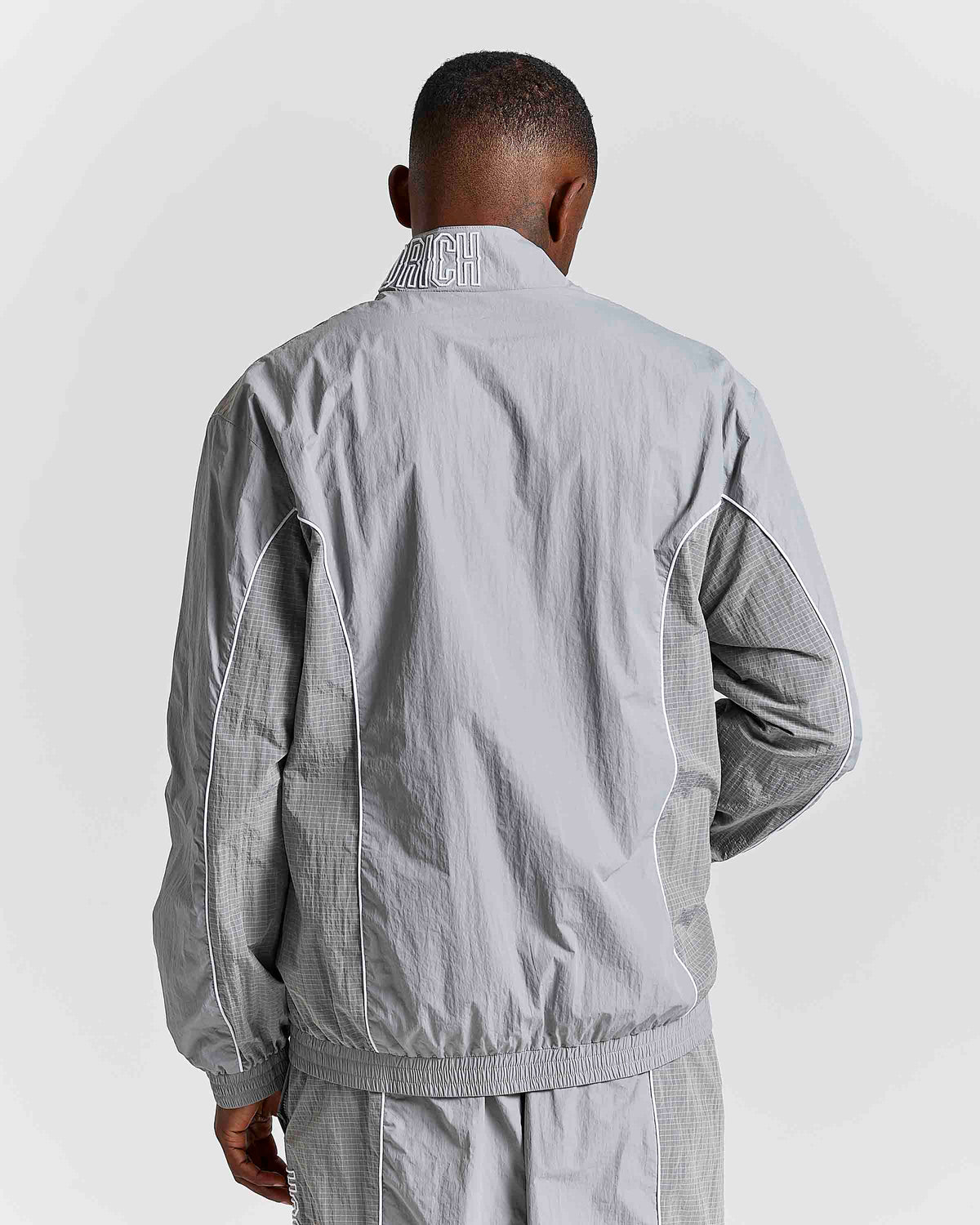 A man viewed from behind wearing a light gray windbreaker jacket with textured gray knit sleeves and a matching gray track pants, against a light gray studio background.