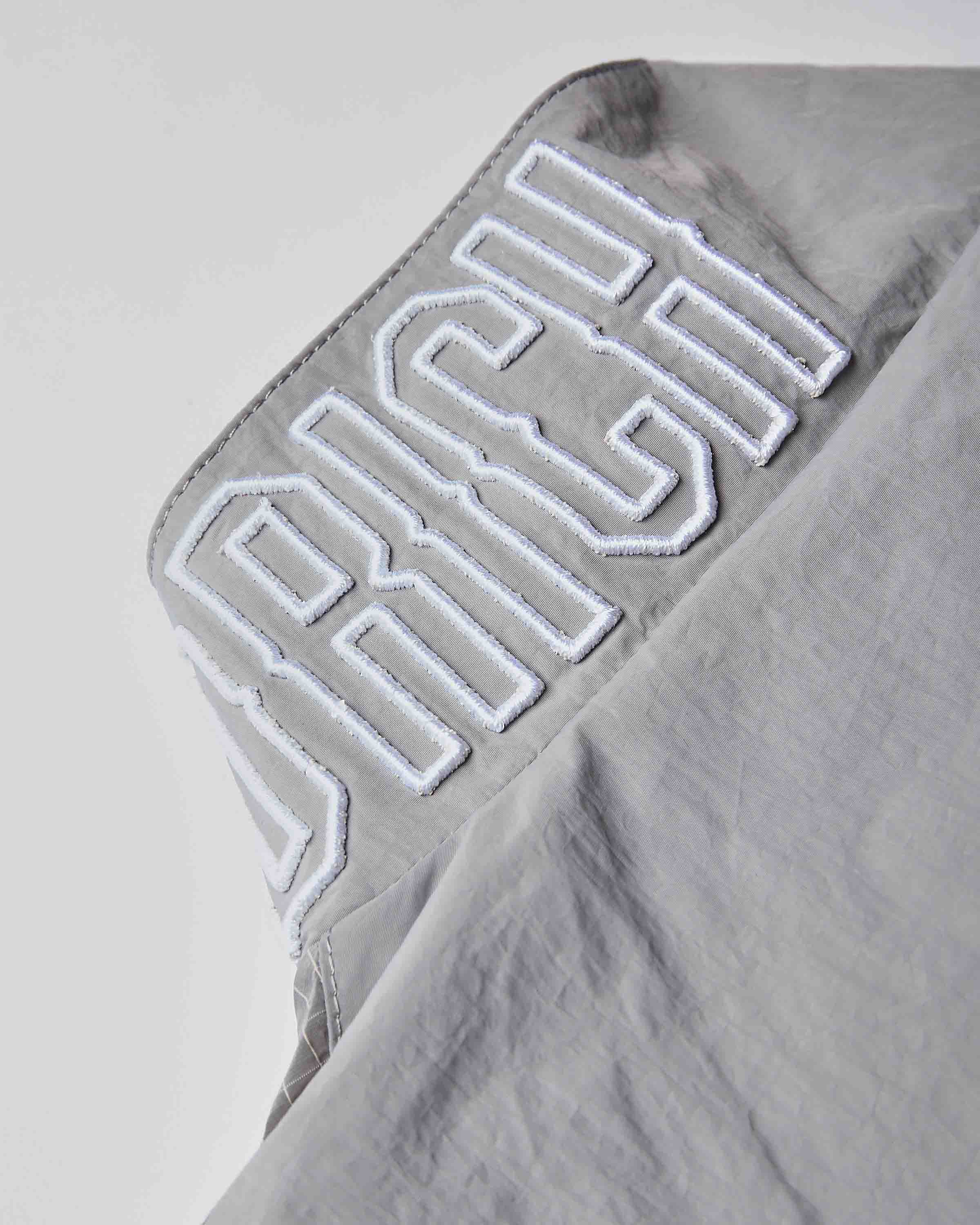 Close-up of a light gray fabric garment with large white embroidered lettering reading 'RICH' on the shoulder.