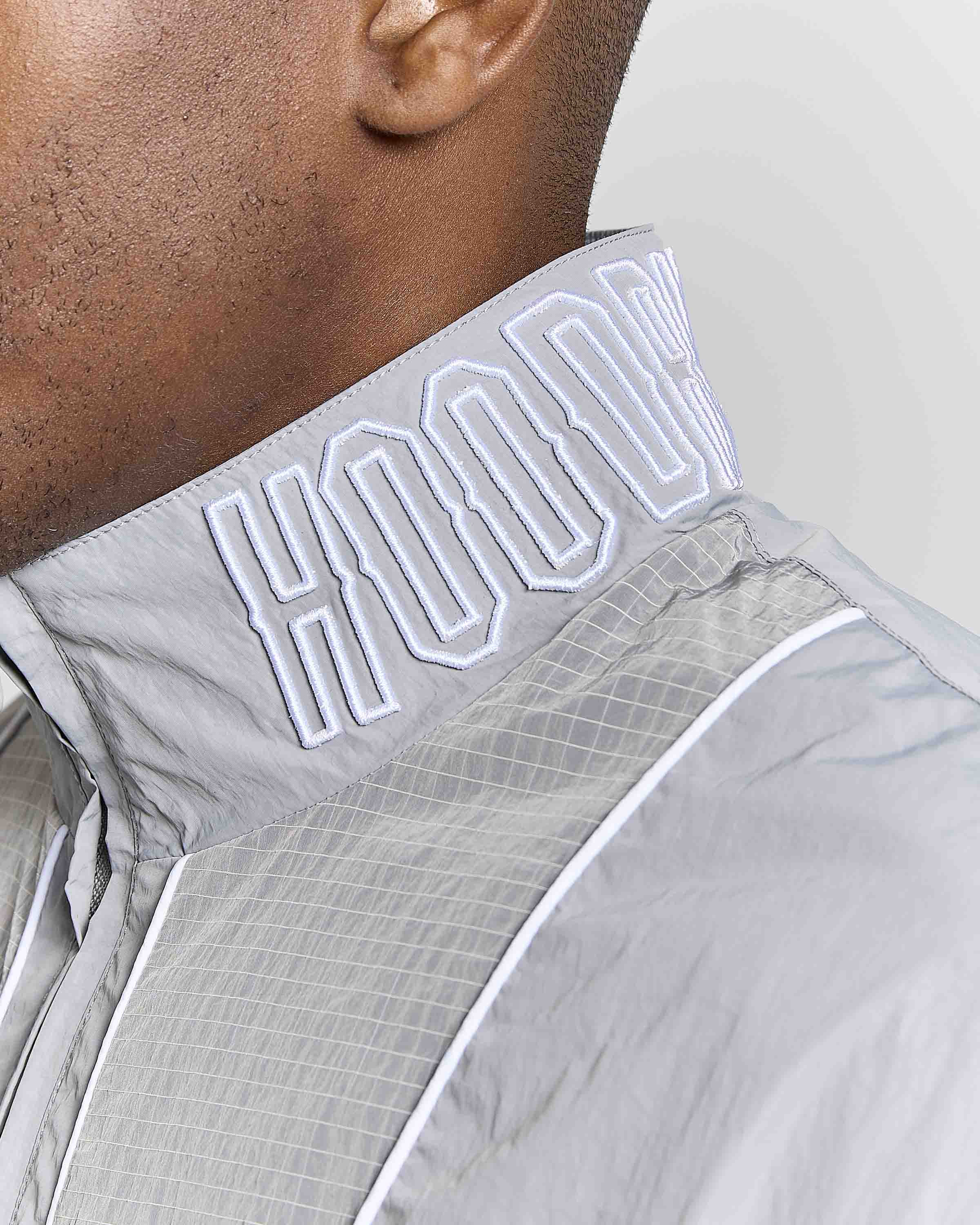 Close-up of a model wearing a light gray hooded windbreaker jacket, highlighting the high collar with white embroidered 'HOODRICH' lettering.