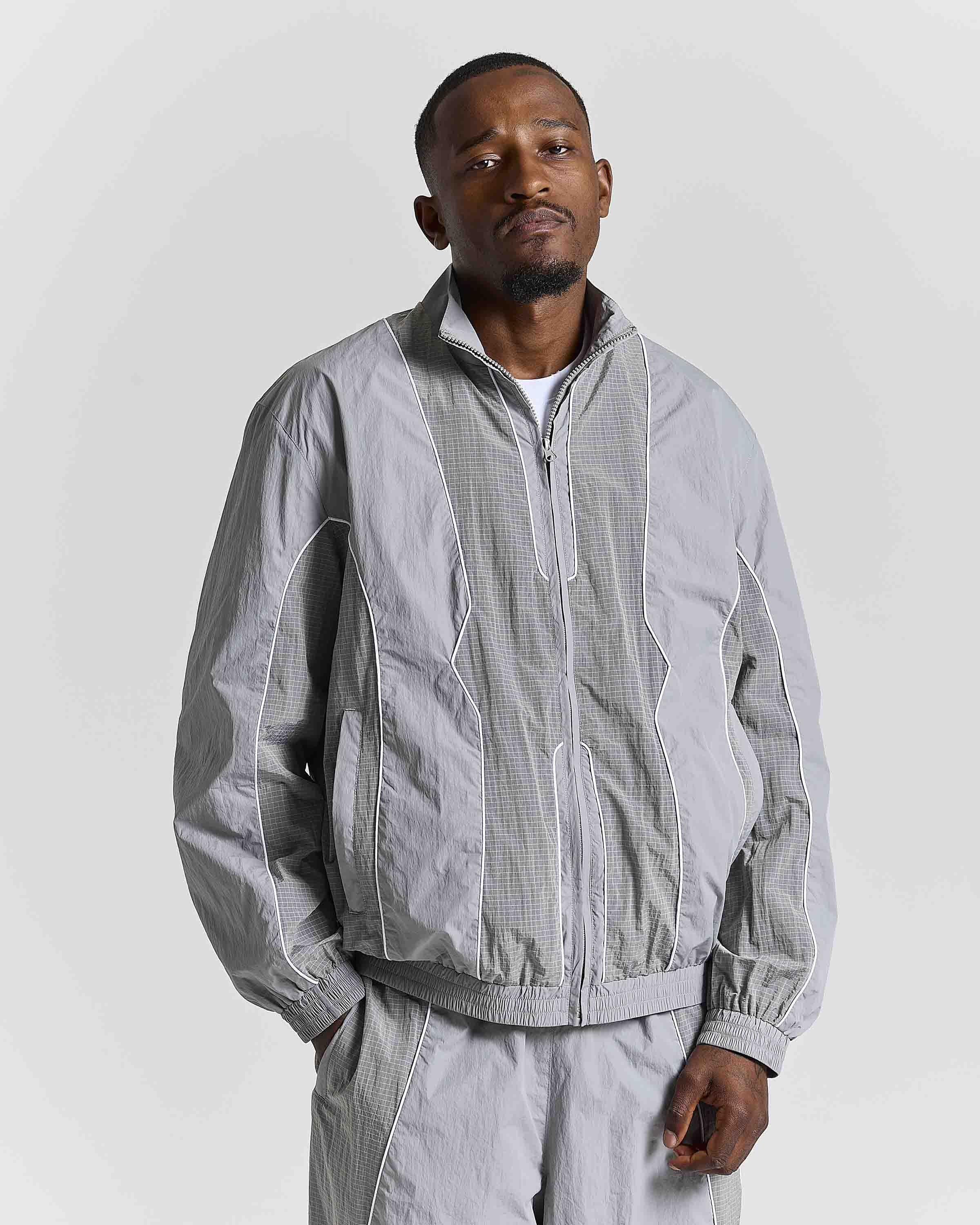 A man wearing a light gray zip-up windbreaker jacket with matching light gray trousers, photographed against a plain light gray studio background.