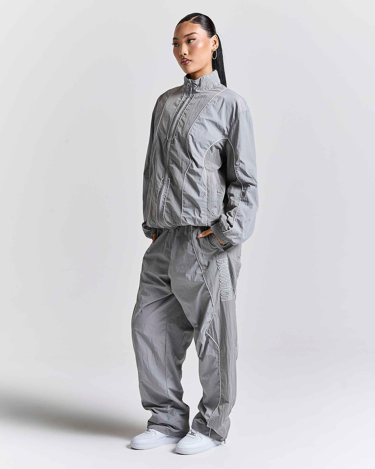 A model wearing a light gray technical windbreaker jacket and matching loose-fit pants, standing against a plain light gray studio background.