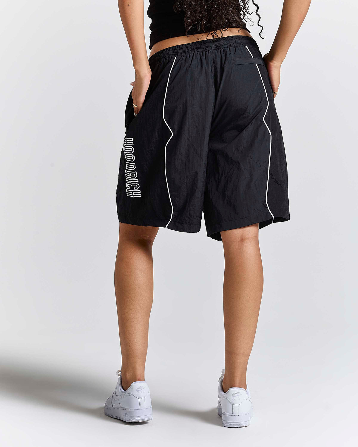 A person shown from the back wearing black athletic shorts with white piping and white sneakers, photographed against a light gray studio background.