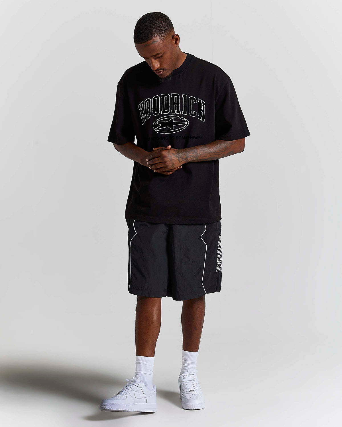 A man modeling a black Hoodrich t-shirt with a white graphic logo, paired with black athletic shorts, white socks, and white sneakers, standing against a light gray studio background.