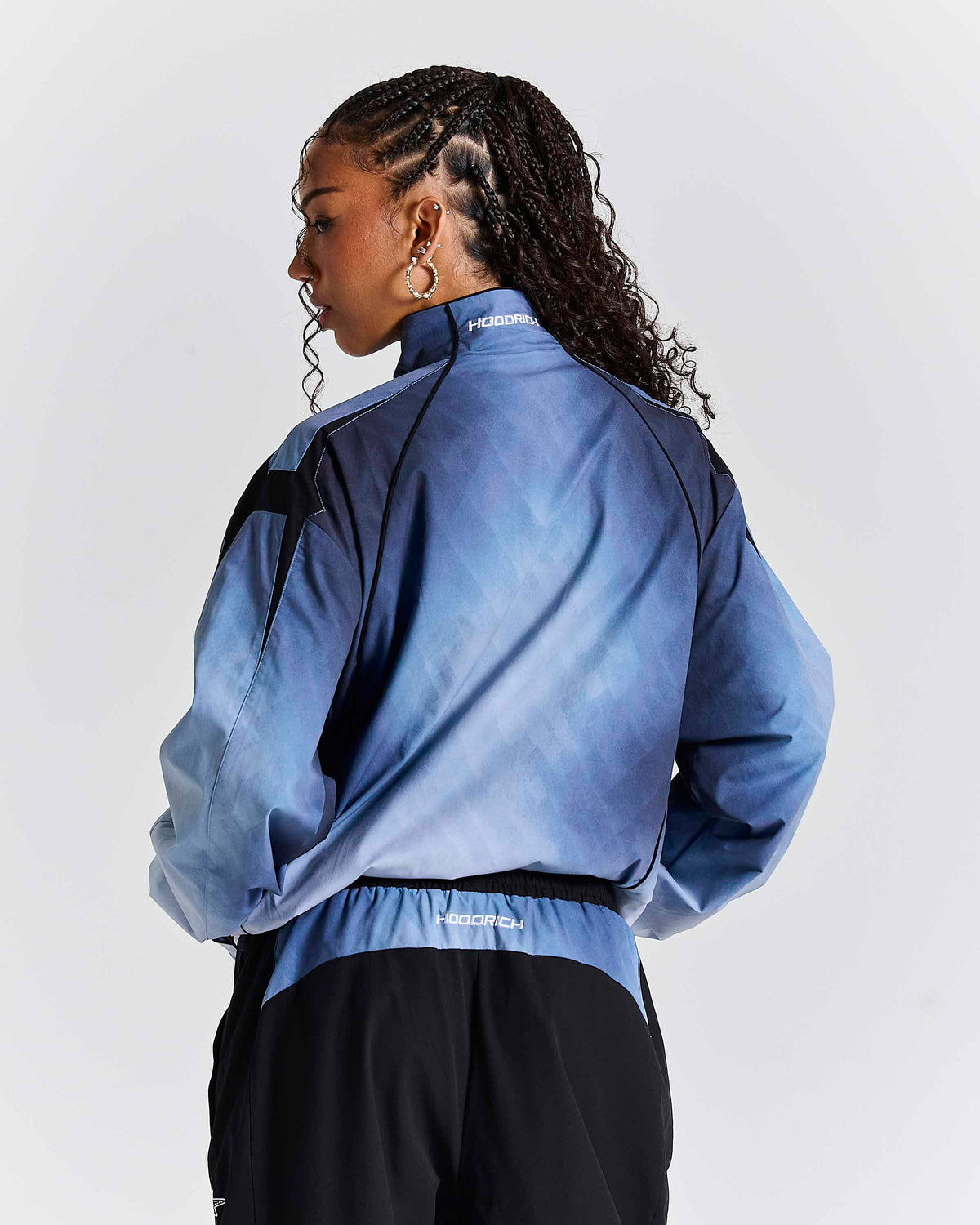 A model shown from the back wearing a blue gradient athletic jacket with black shoulder panels and black track pants against a light gray studio background.