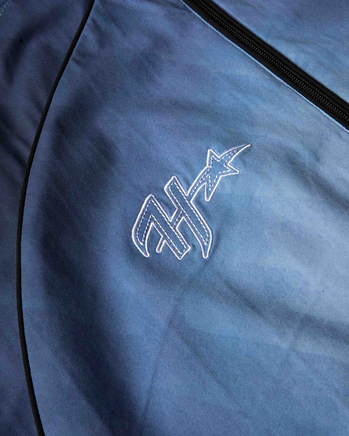 Close-up of a blue zippered garment or bag with black piping and a white embroidered stylized logo featuring a star.