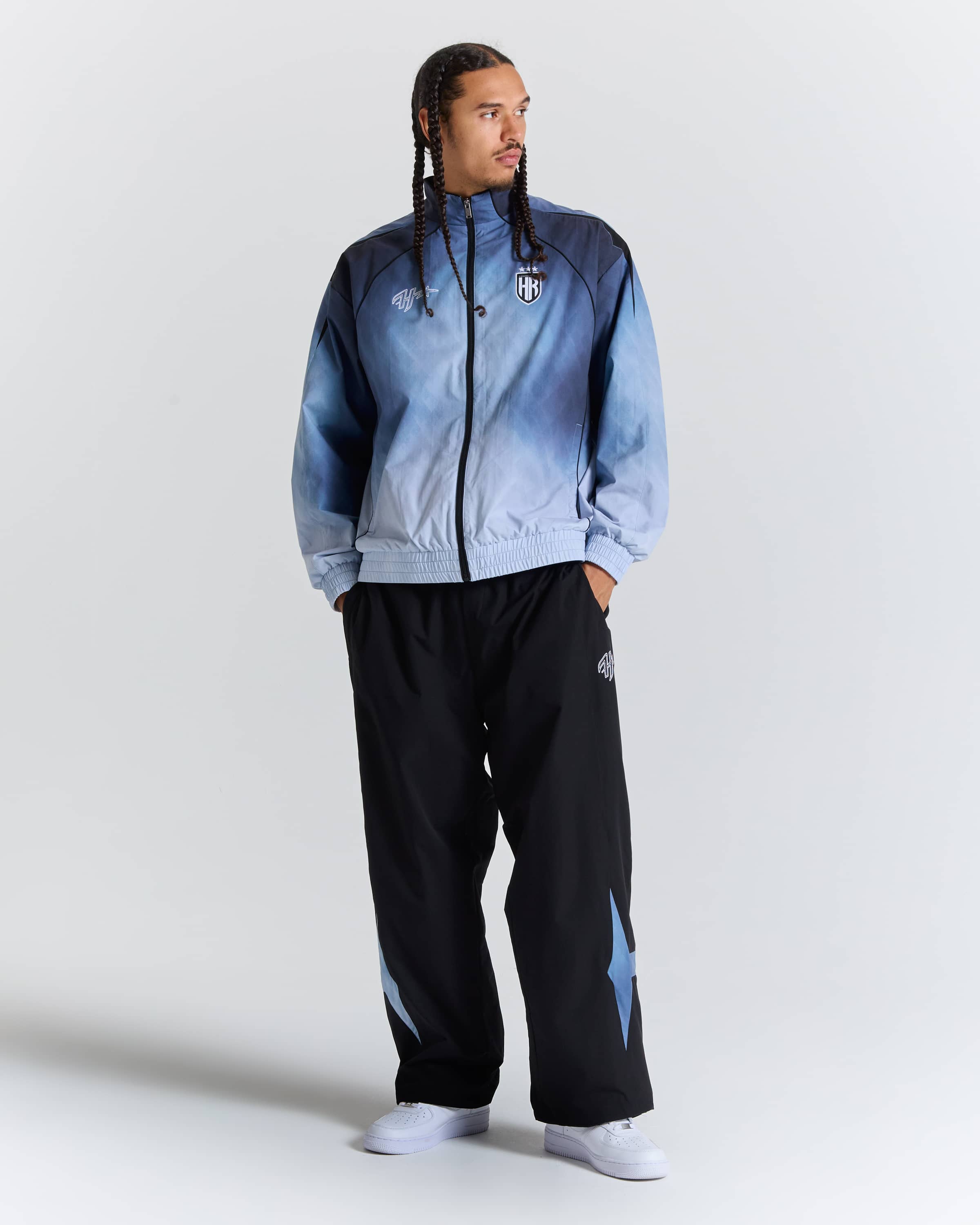 A man modeling a light blue and navy gradient zip-up jacket with black track pants featuring light blue star details, set against a plain light gray studio background.