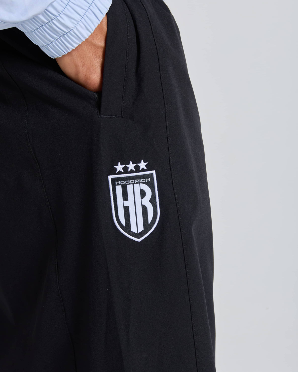 Close-up of a person wearing black hooded sweatshirt or jacket with a white embroidered shield logo featuring the letters HR and the word HOODRICH above it, with three stars at the top. A light blue ribbed sleeve is also visible at the top left.