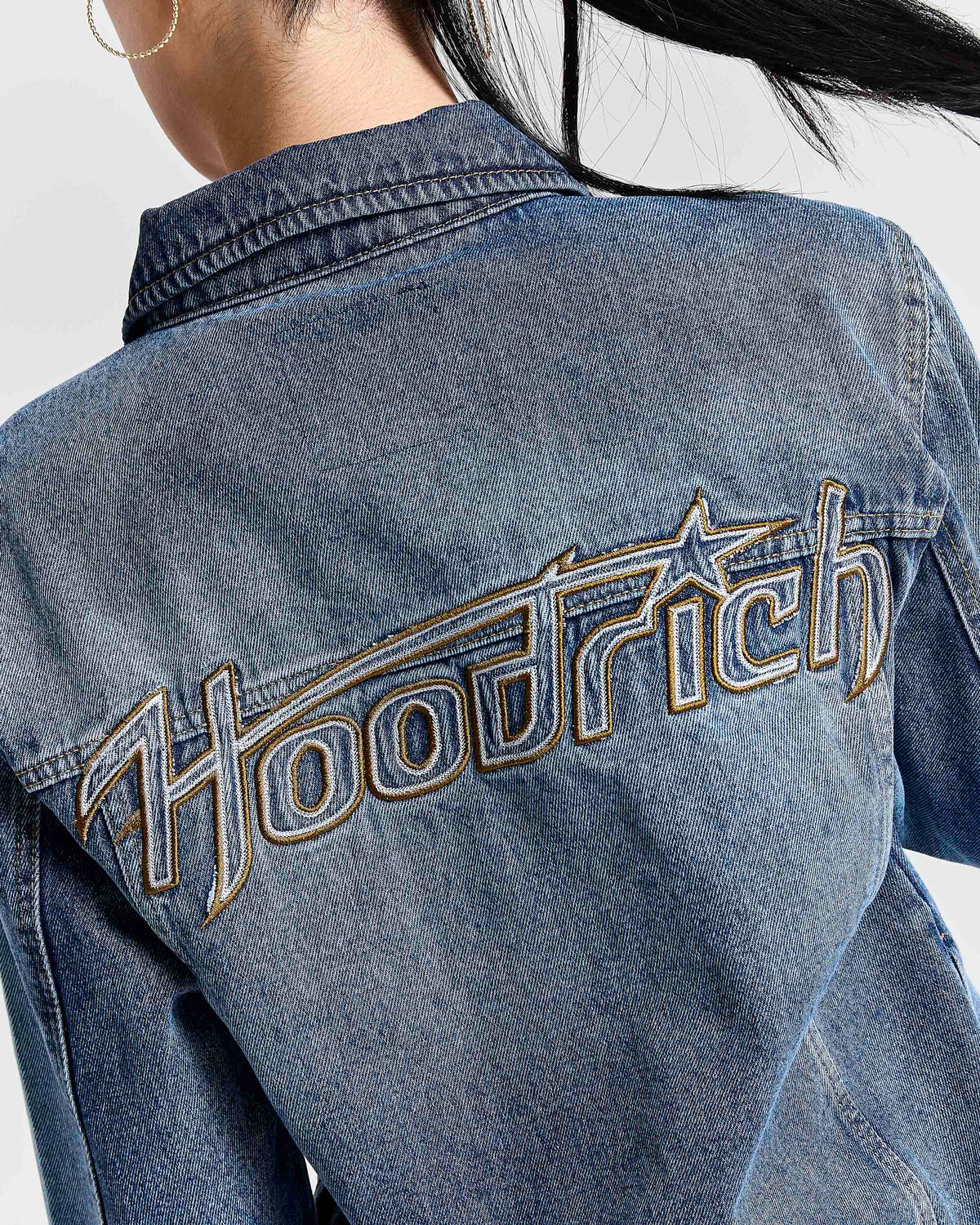 Close-up back view of a person wearing a blue denim jacket with large metallic embroidered 'Hoodrich' lettering across the upper back.
