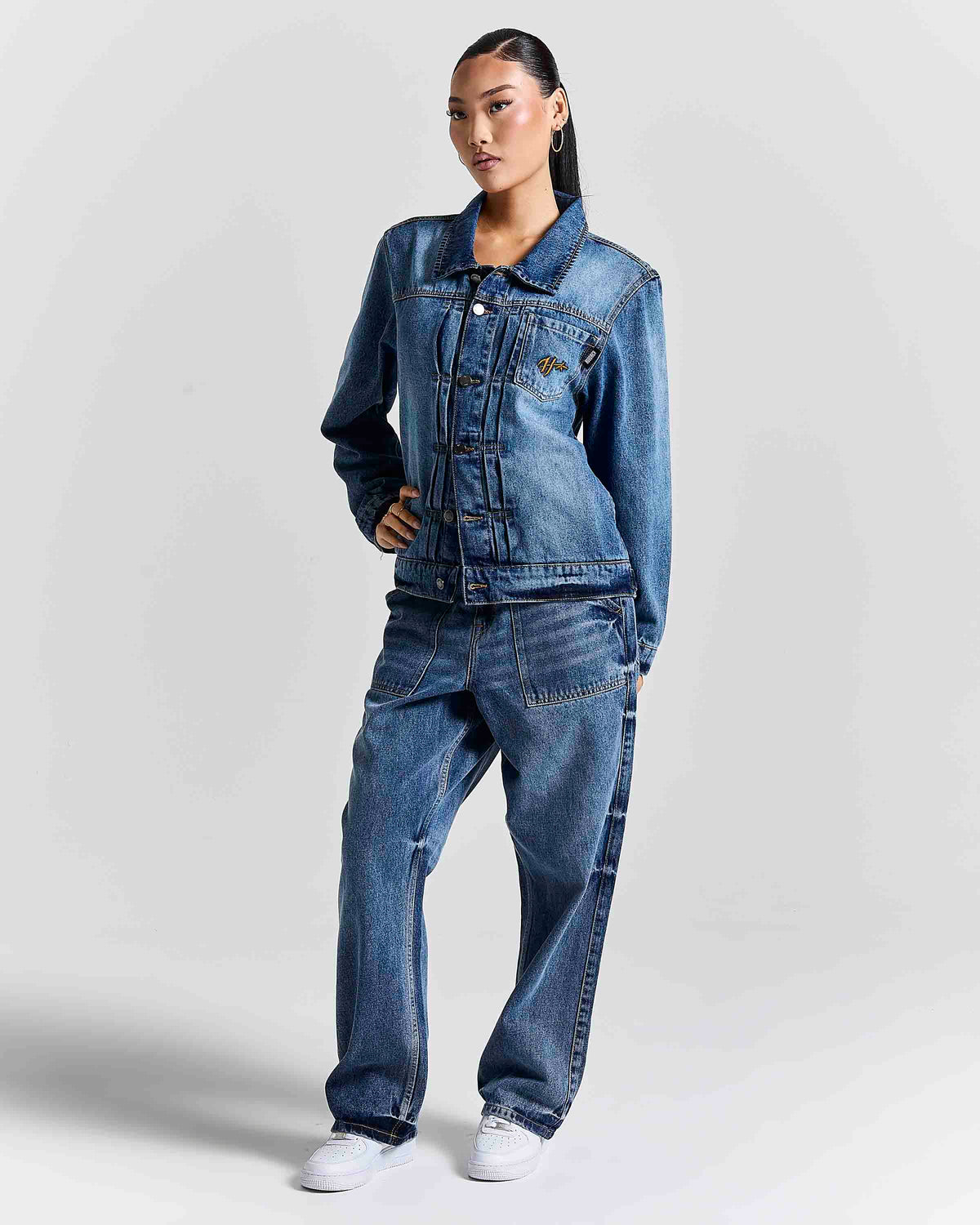 A model wearing a matching blue denim jacket and wide-leg denim jeans set, paired with white sneakers, standing against a light gray studio background.