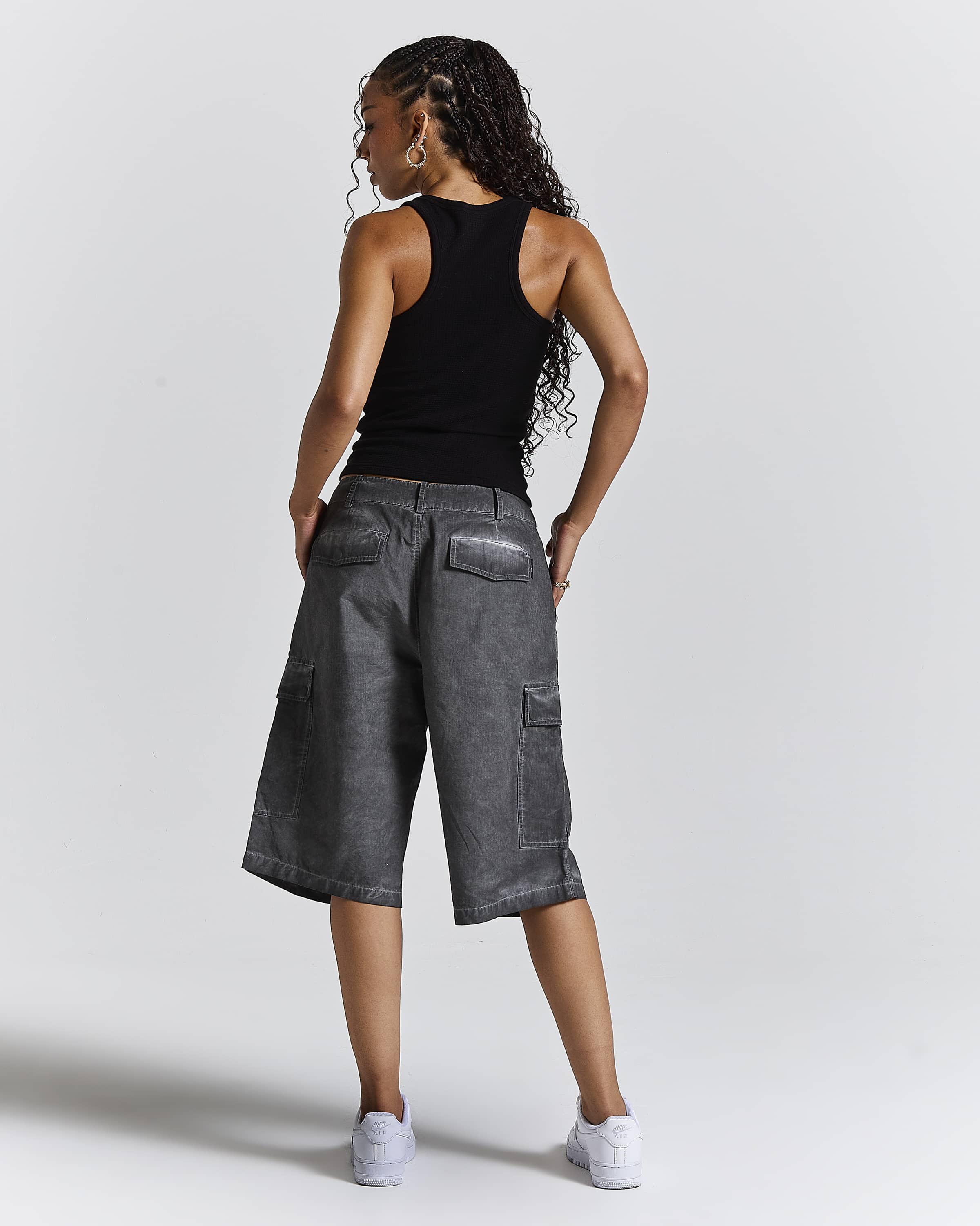 A woman shown from the back wearing washed charcoal gray cropped cargo shorts, a black tank top, and white sneakers against a light gray studio background.