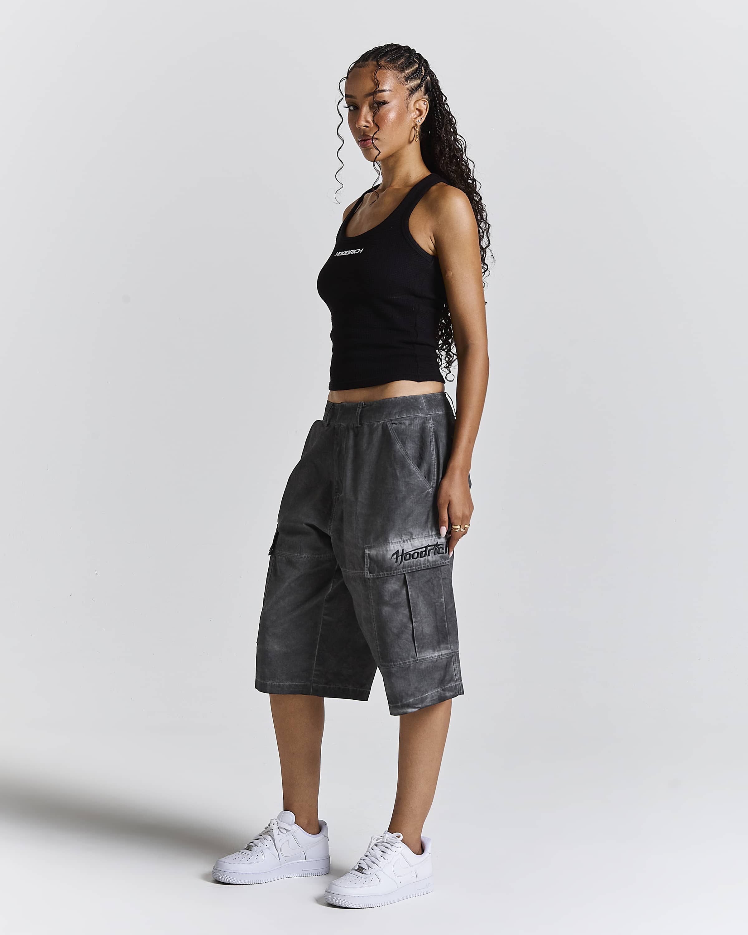 A woman modeling dark gray washed cargo shorts with a white script logo on the pocket, paired with a black tank top and white sneakers, against a light gray studio background.