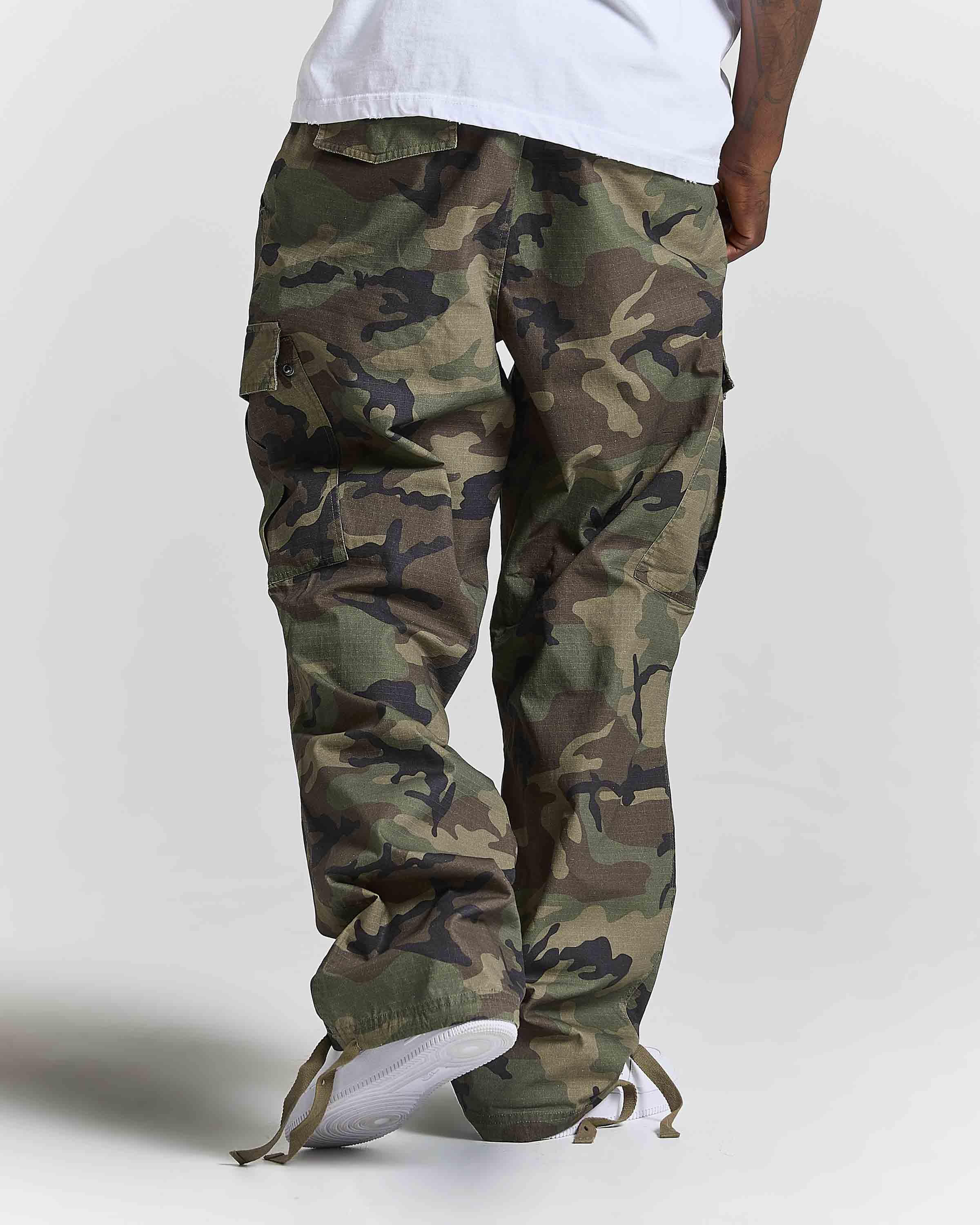 A person shown from the waist down wearing loose-fit camouflage cargo pants with a white t-shirt and white sneakers, photographed against a light gray studio background.