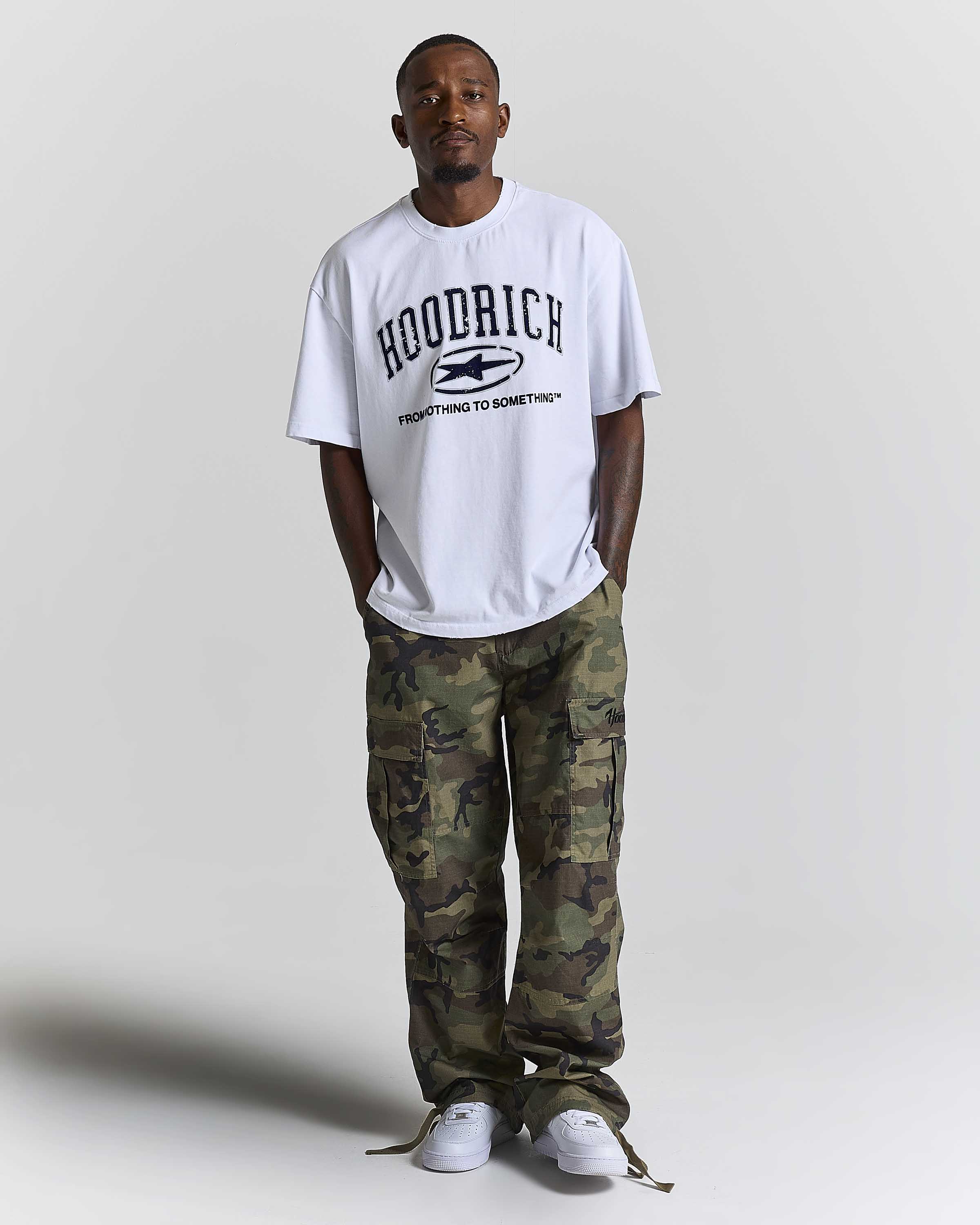 A man wearing a white HOODRICH t-shirt with a black graphic print, paired with green and brown camouflage cargo pants and white sneakers, standing against a light gray studio background.