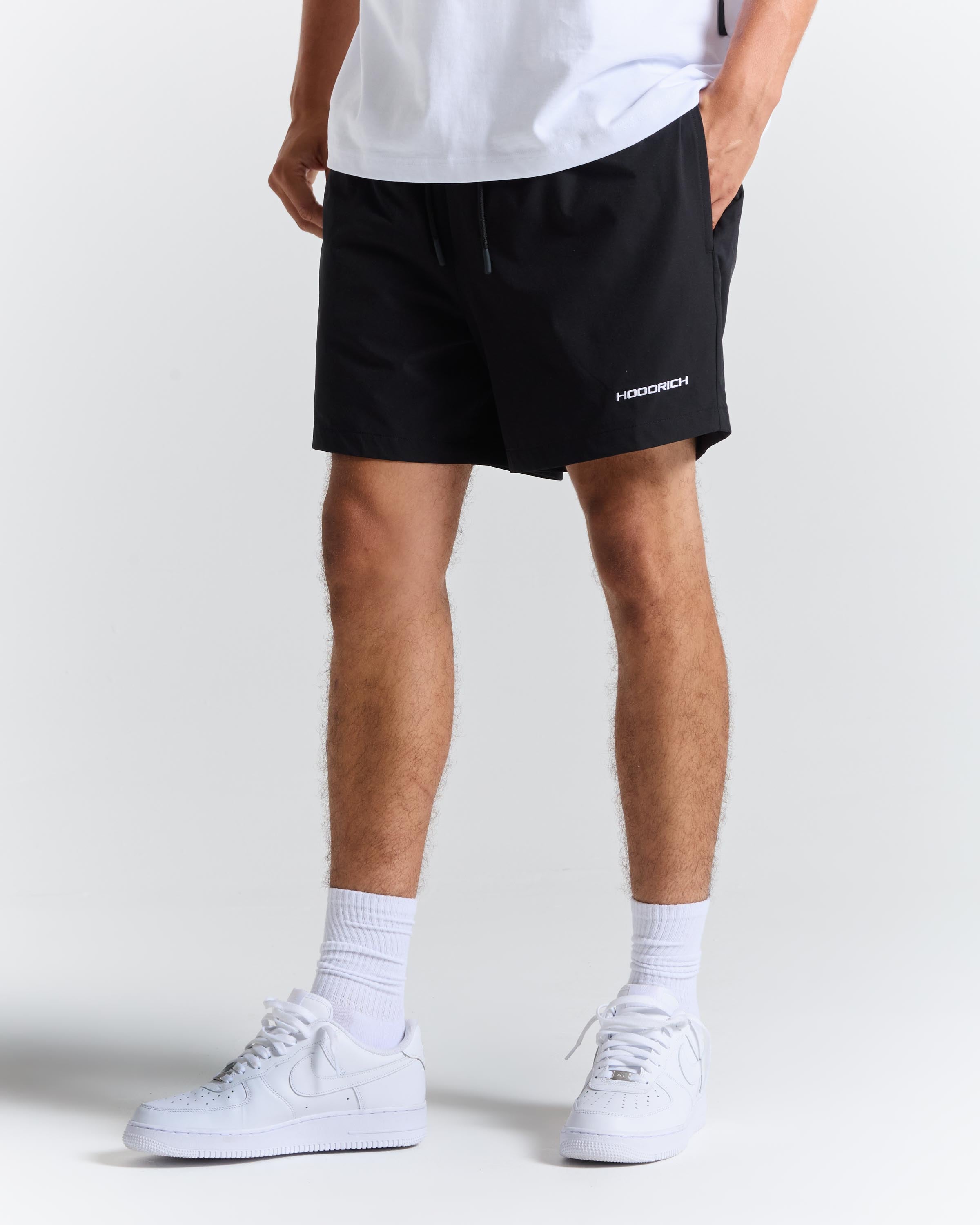 A person wearing black athletic shorts with a small white 'HOODRICH' logo on the lower left leg, paired with a white t-shirt, white socks, and white sneakers, shown from the waist down on a light gray studio background.