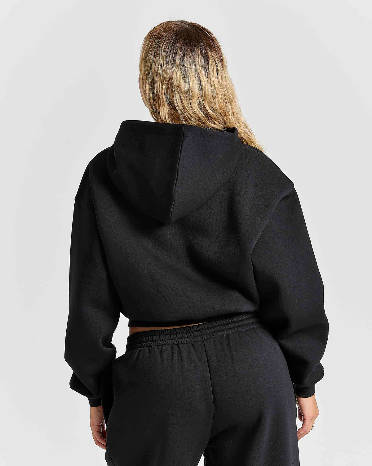 A woman viewed from behind wearing a black cropped hoodie and black sweatpants against a light gray studio background.