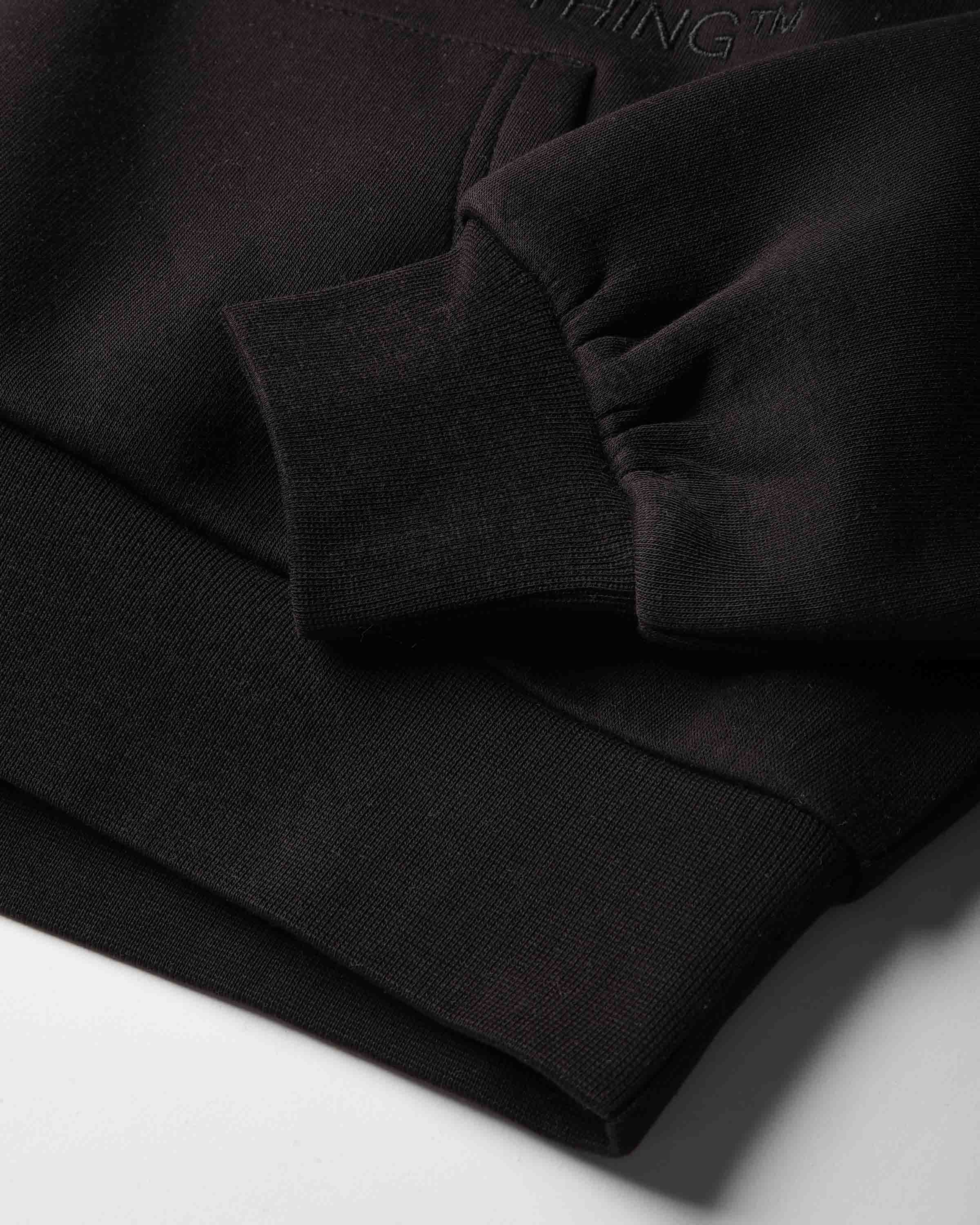 Close-up of a black ribbed knit garment, likely a sweatshirt or hoodie, showing the cuff, hem, and a small tonal embroidered brand mark reading NOTHING on a white surface.
