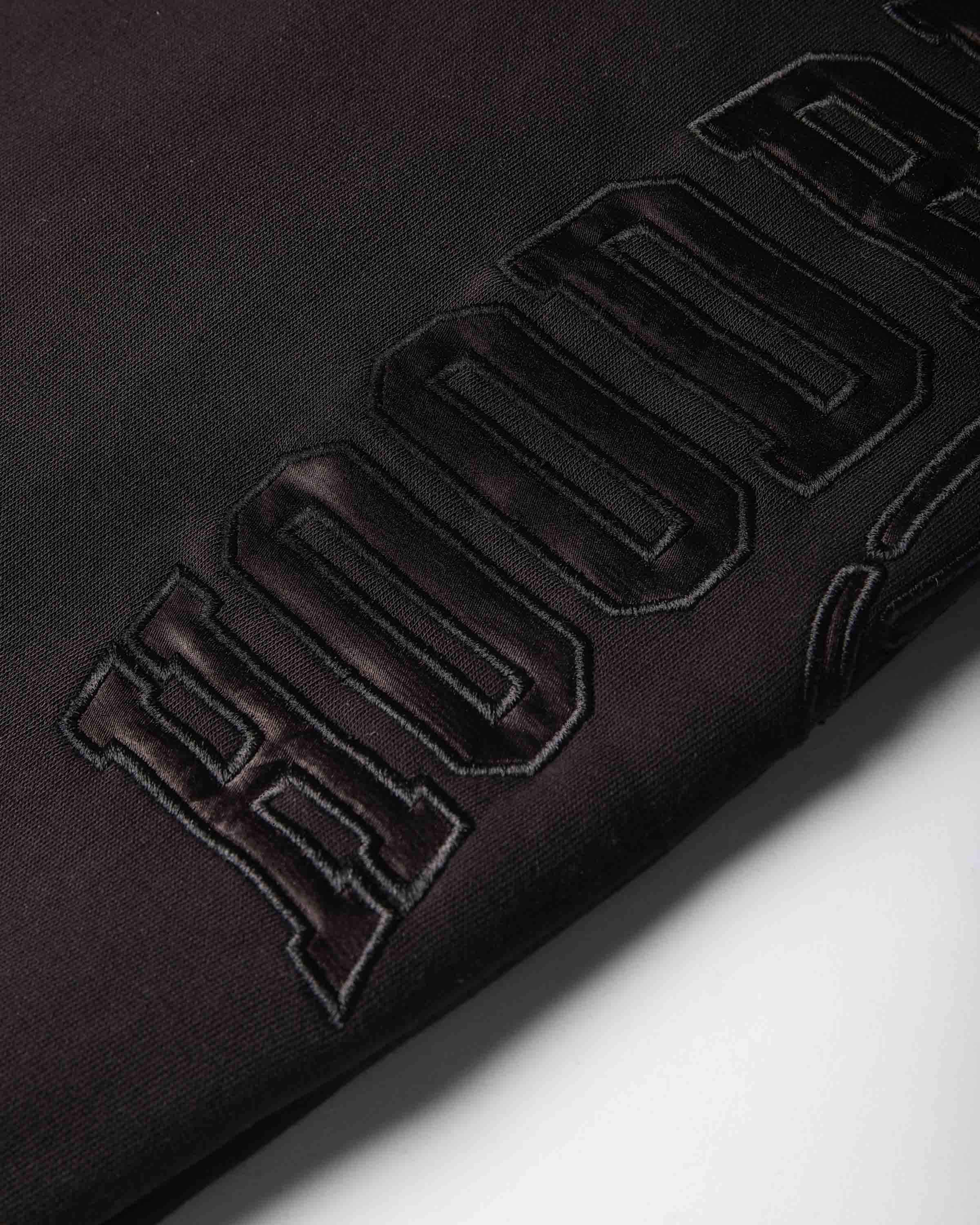 Close-up of a black fabric surface with large raised embroidered lettering in a dark metallic burgundy tone, photographed on a white background.