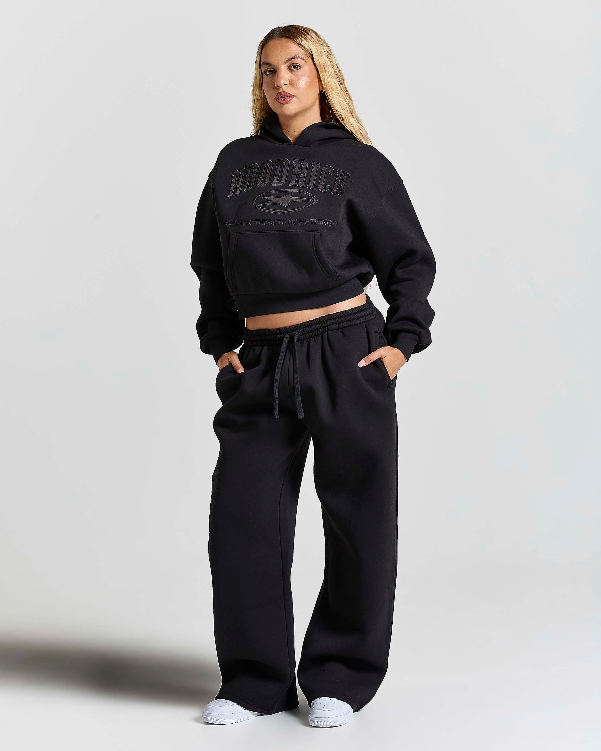 A woman modeling a black cropped hoodie and matching wide-leg sweatpants set. The hoodie features a tonal black graphic on the chest with the word 'HOODRICH' on the front.