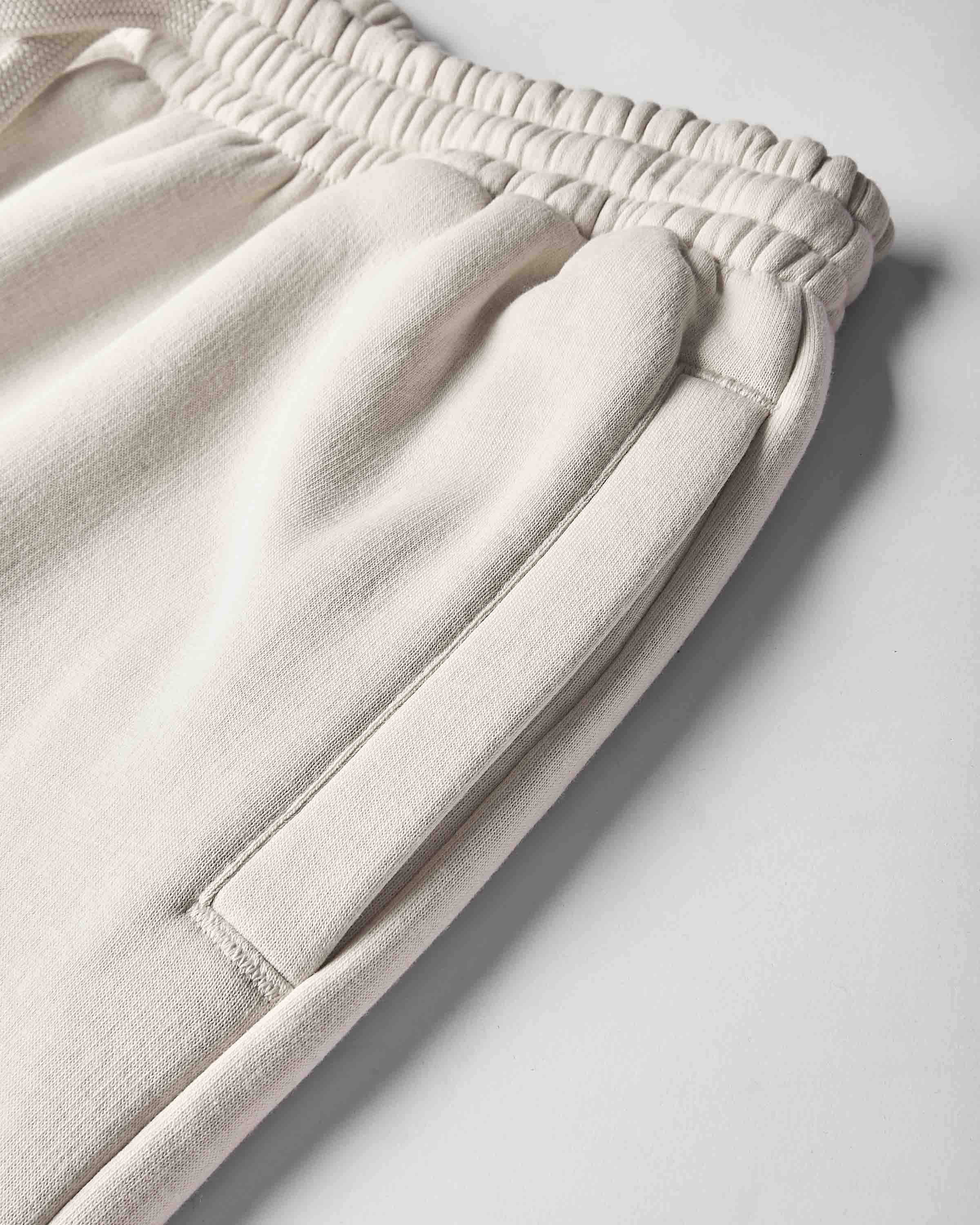 Close-up of a pair of light cream or off-white sweatpants showing the elastic waistband, side pocket seam, and soft fabric texture on a light gray background.