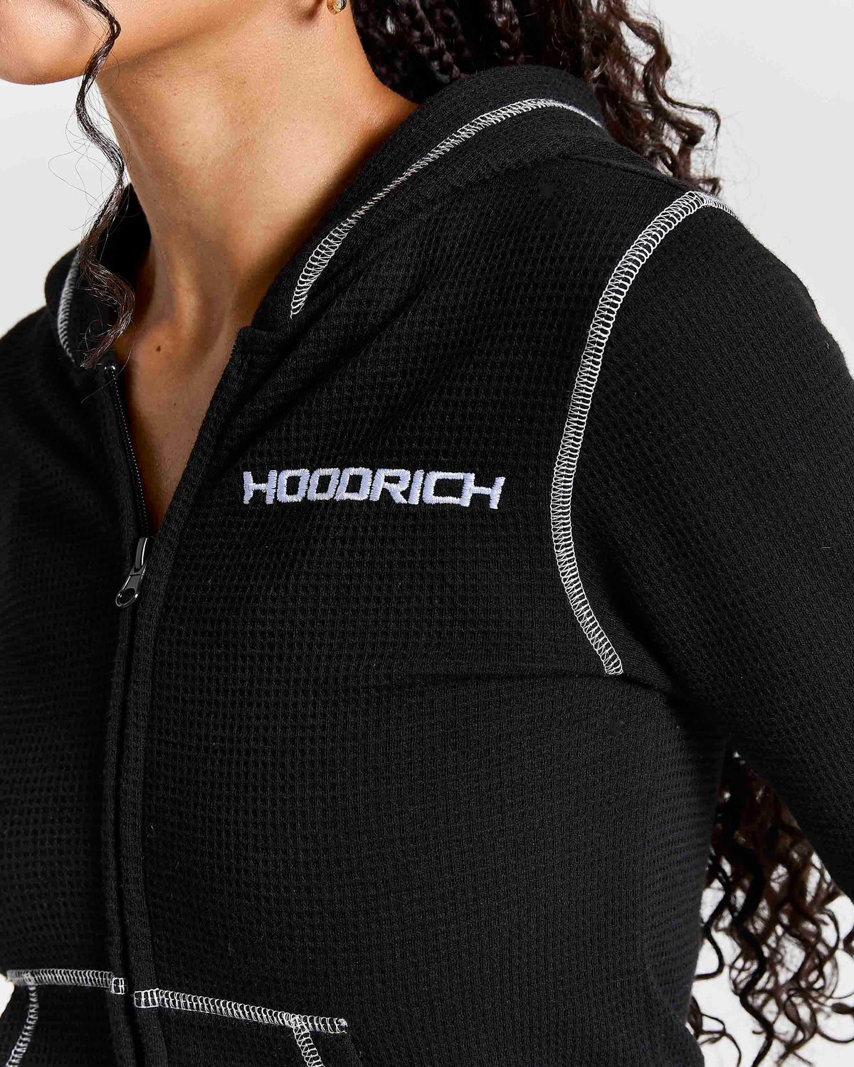 Close-up of a person wearing a black waffle-knit zip-up hoodie with white contrast stitching and a white HOODRICH logo on the chest.