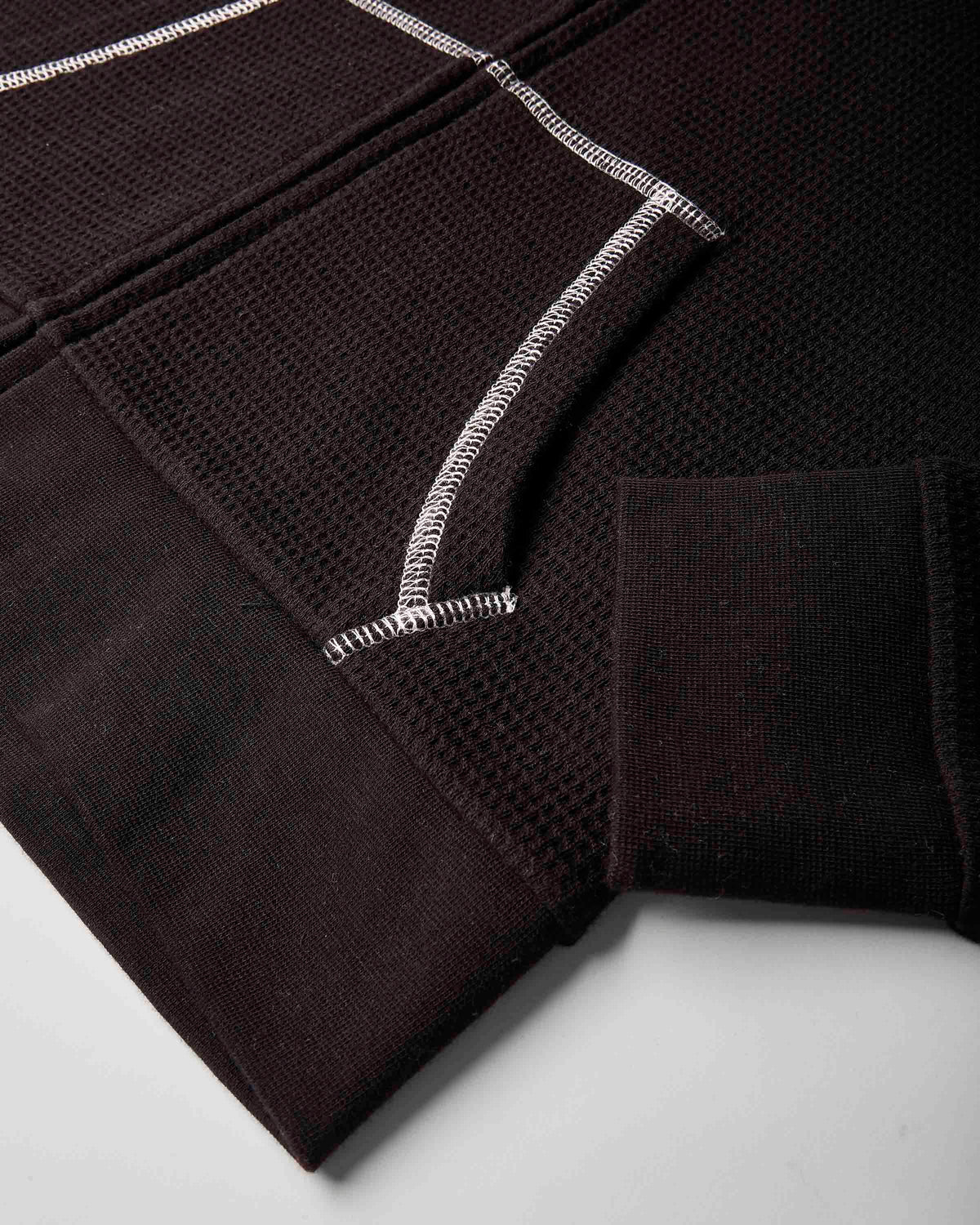 Close-up of a dark brown textured knit garment, showing a ribbed cuff, a waffle-knit panel, and contrasting white zigzag stitching on a light gray background.