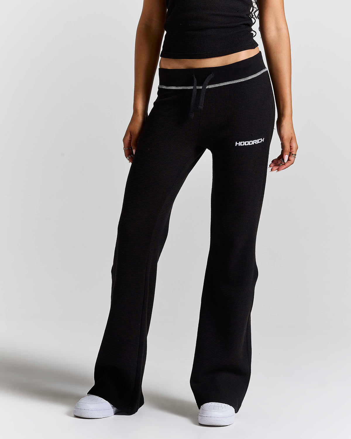 A model wearing black wide-leg sweatpants with a white drawstring and a white 'HOODRICH' logo on the left thigh, paired with a black crop top and white sneakers, set against a plain light gray studio background.