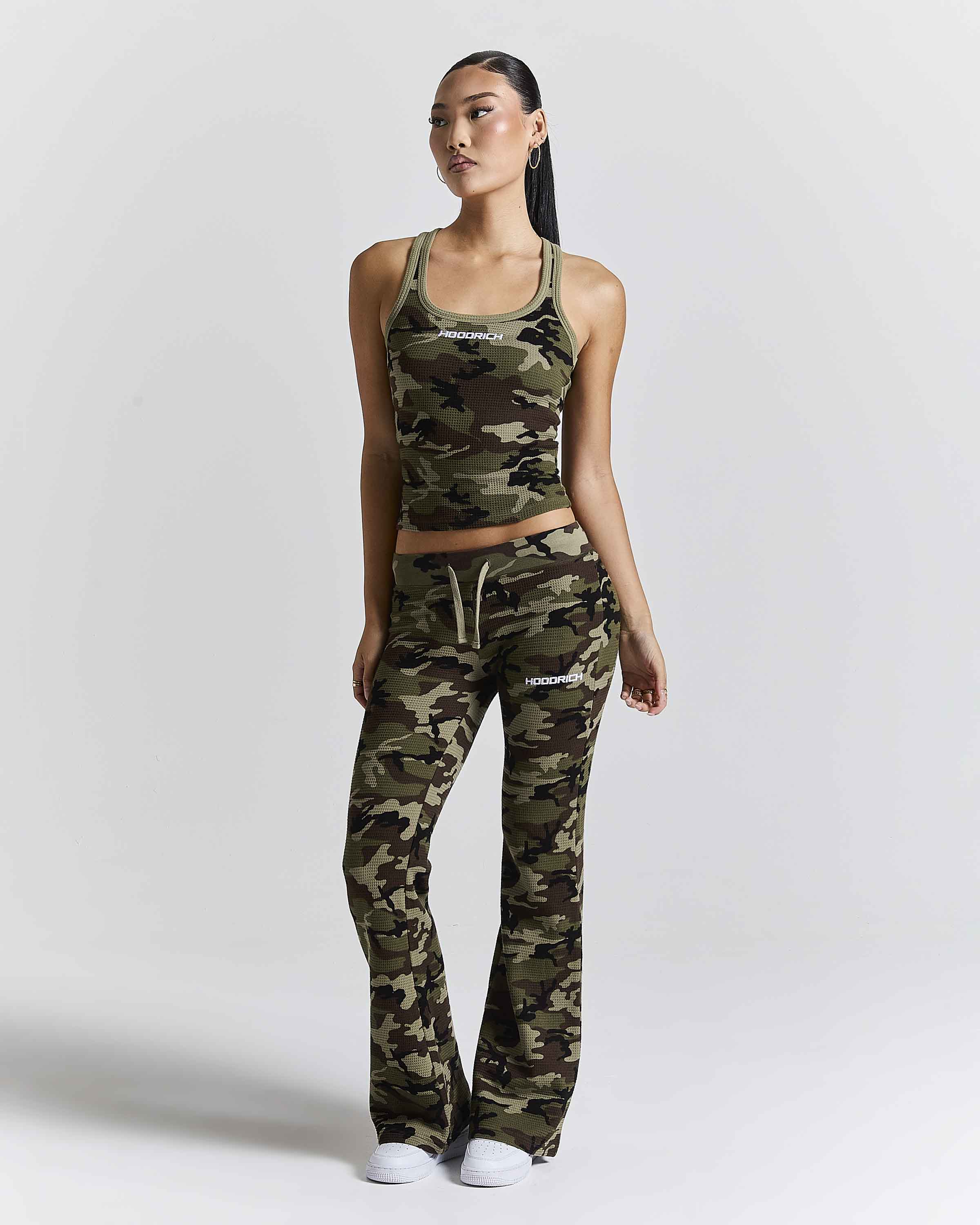A woman modeling a matching camouflage athletic set consisting of a cropped tank top and high-waisted flared pants, photographed against a light gray studio background.