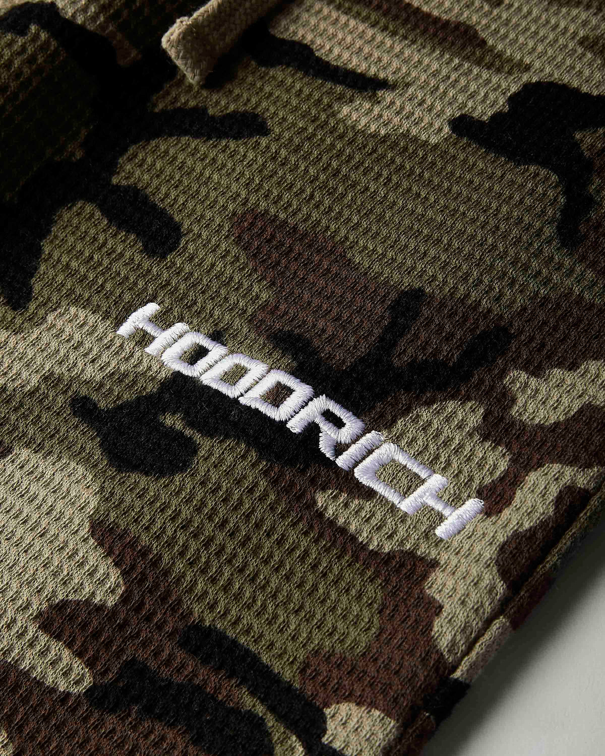 Close-up of a camouflage knit garment with a white embroidered 'HOODRICH' logo on the chest.