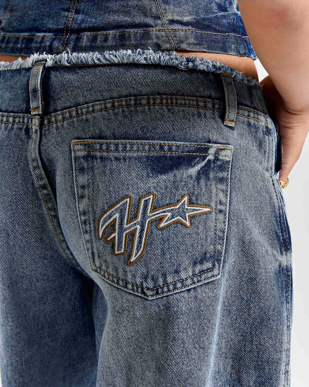 Close-up back view of a person wearing blue denim jeans with a frayed waistband and a large embroidered monogram logo on the back pocket.