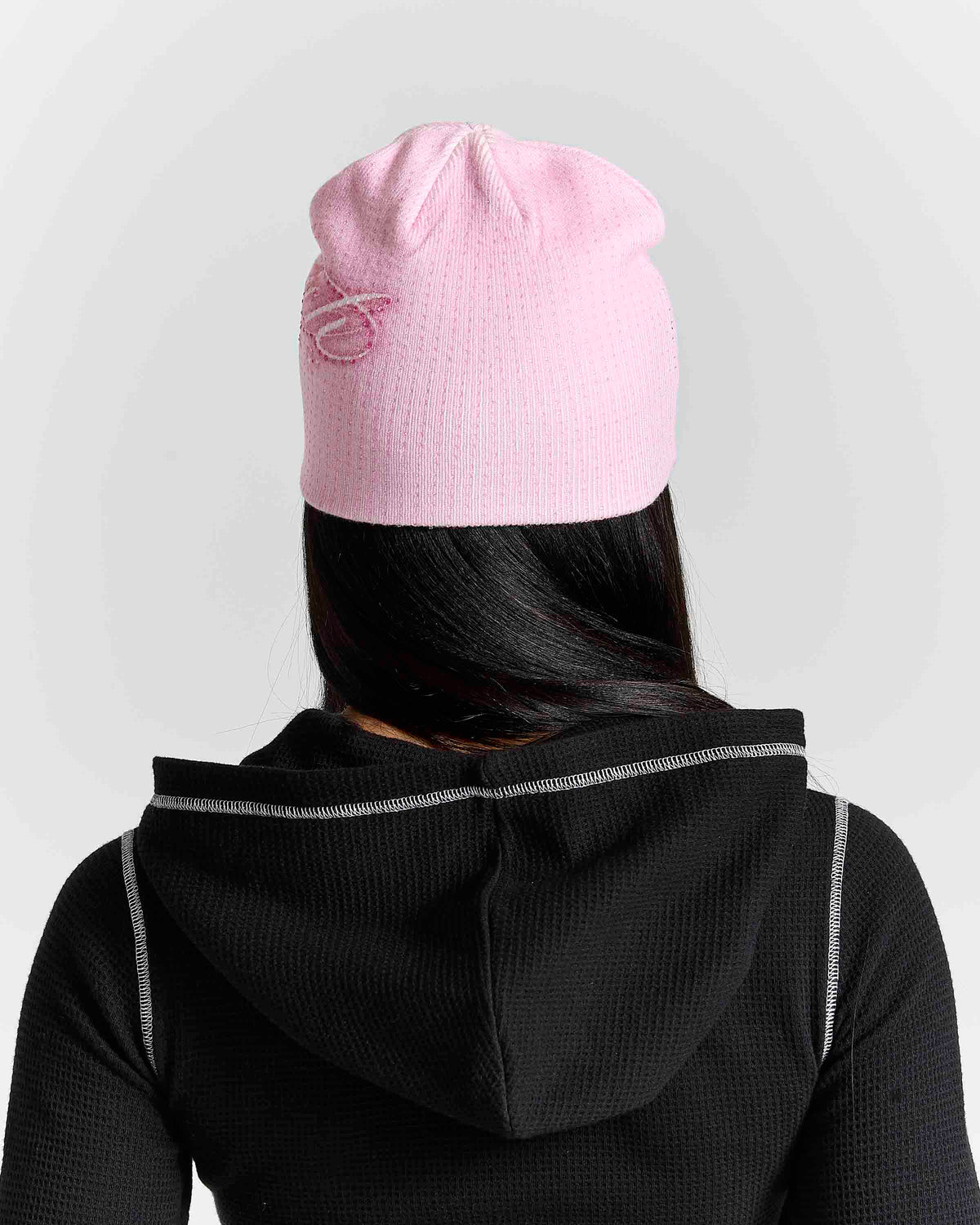 A person shown from the back wearing a light pink knit beanie with a small pink embroidered logo and a black hooded top, against a light gray studio background.