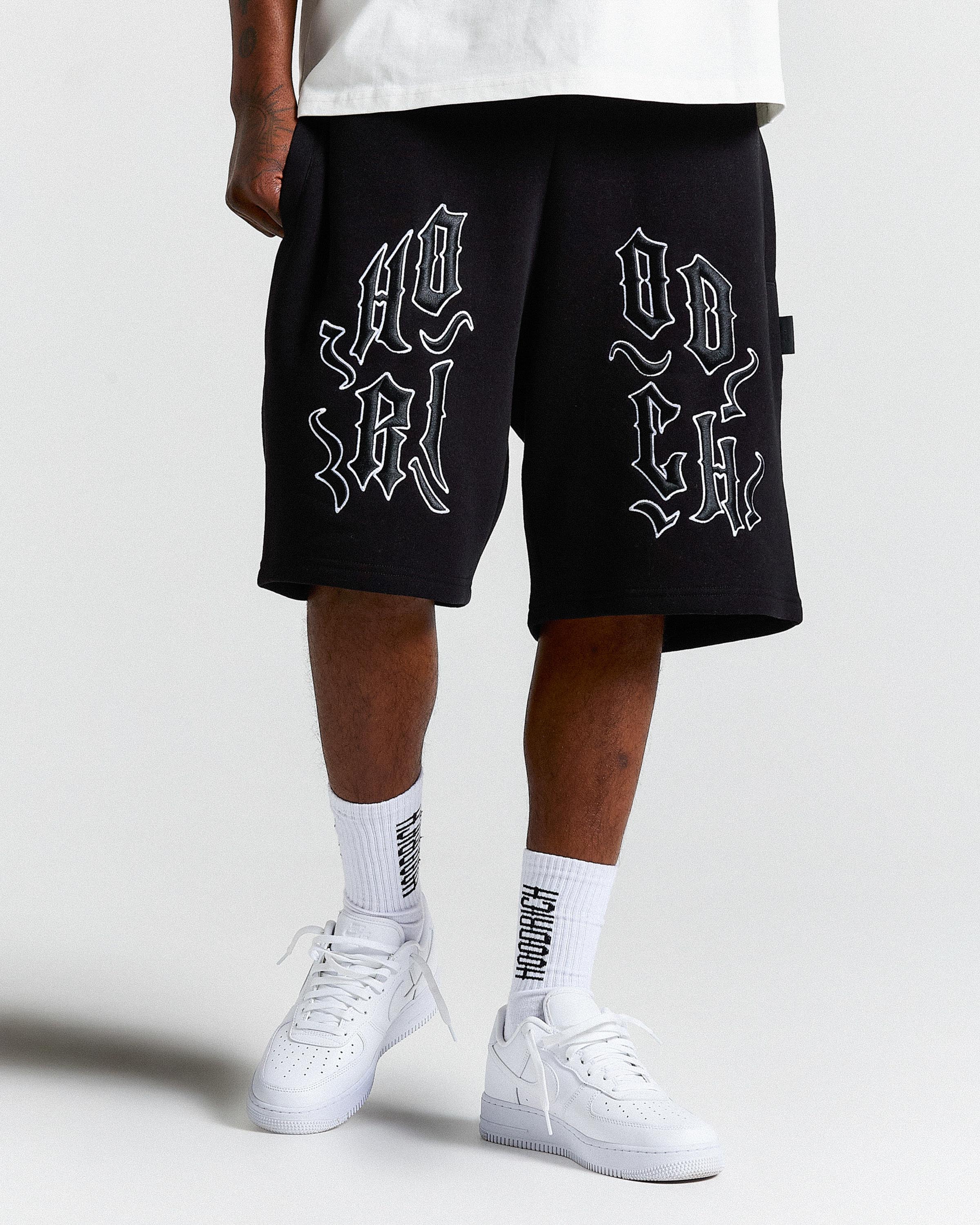 Person wearing black shorts with white text and white sneakers on a light gray background