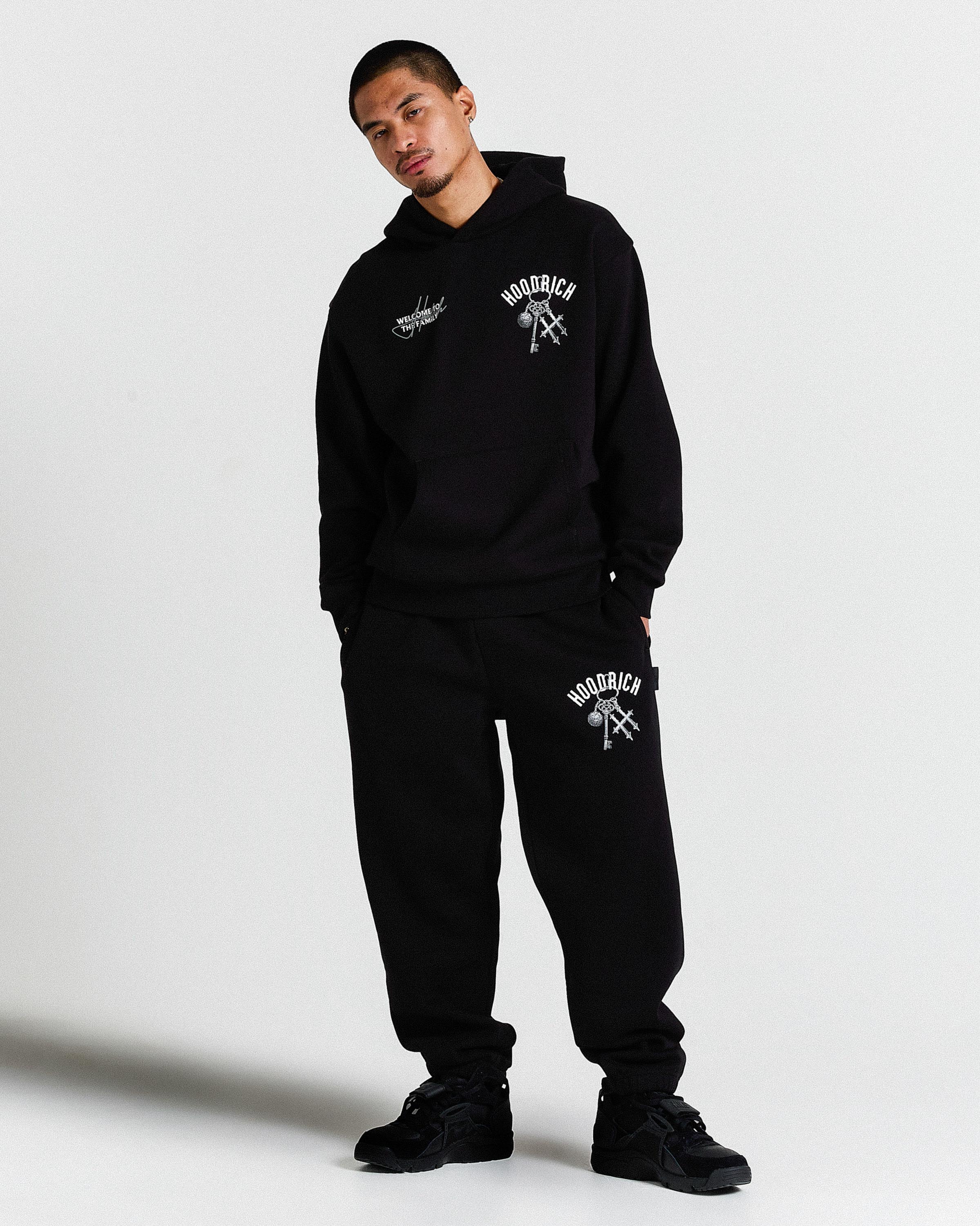 Person wearing a black hoodie and pants with branding on a white background