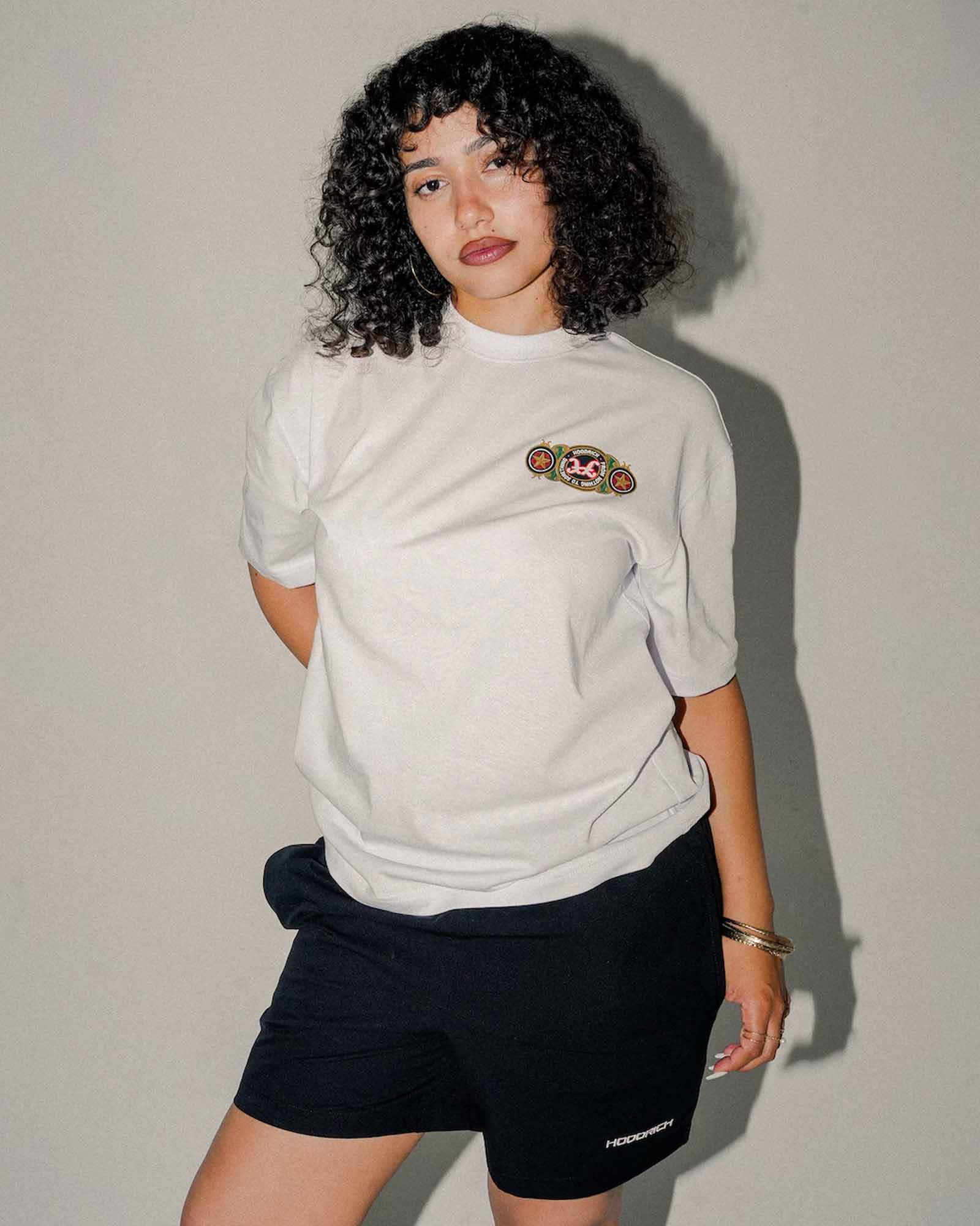 A model with curly black hair wearing a white short-sleeve t-shirt with a colorful graphic on the left chest and black athletic shorts, standing against a plain gray wall.