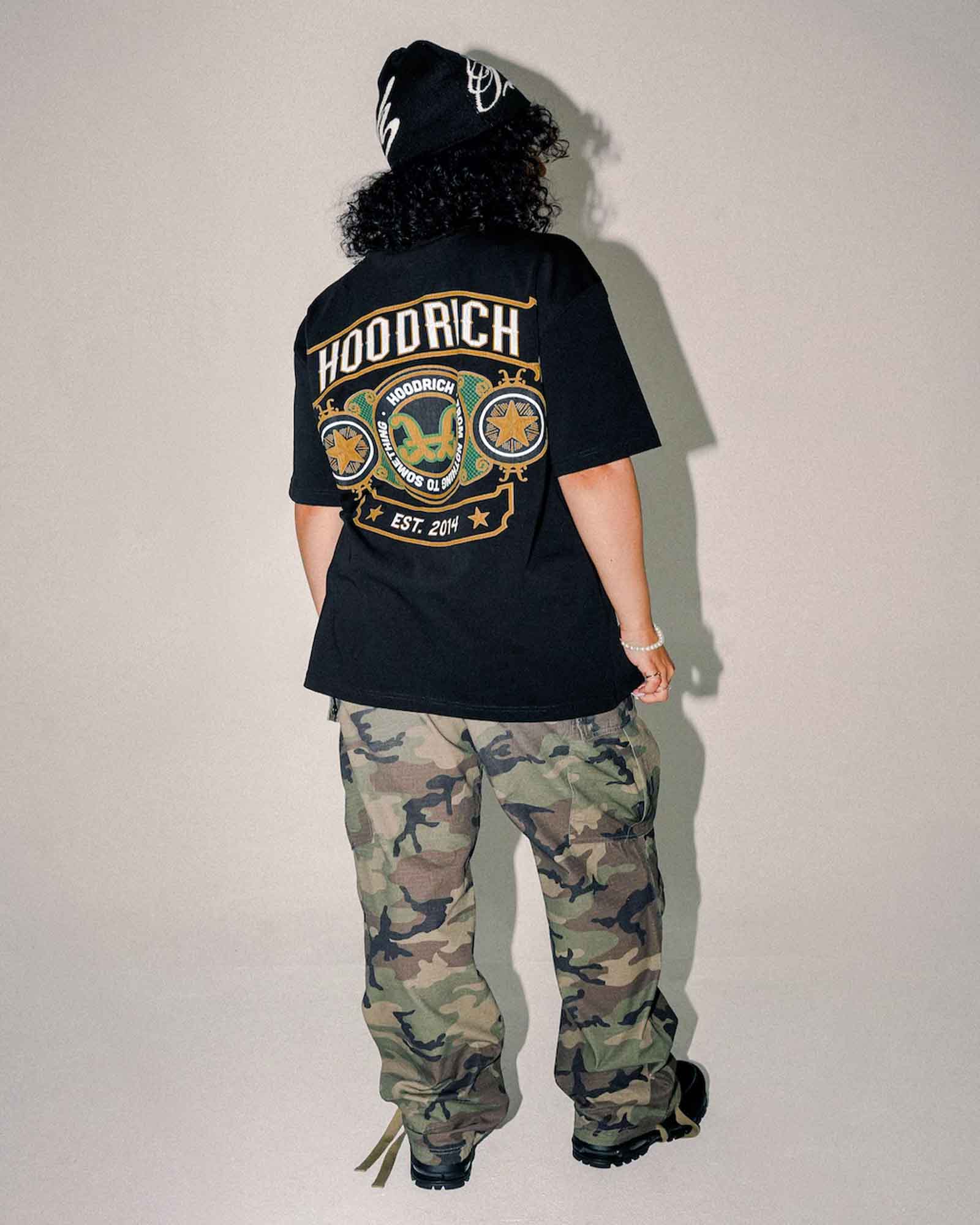 A person shown from the back wearing a black HOODRICH t-shirt with a large graphic print, camouflage pants, and a black beanie against a plain light gray wall.