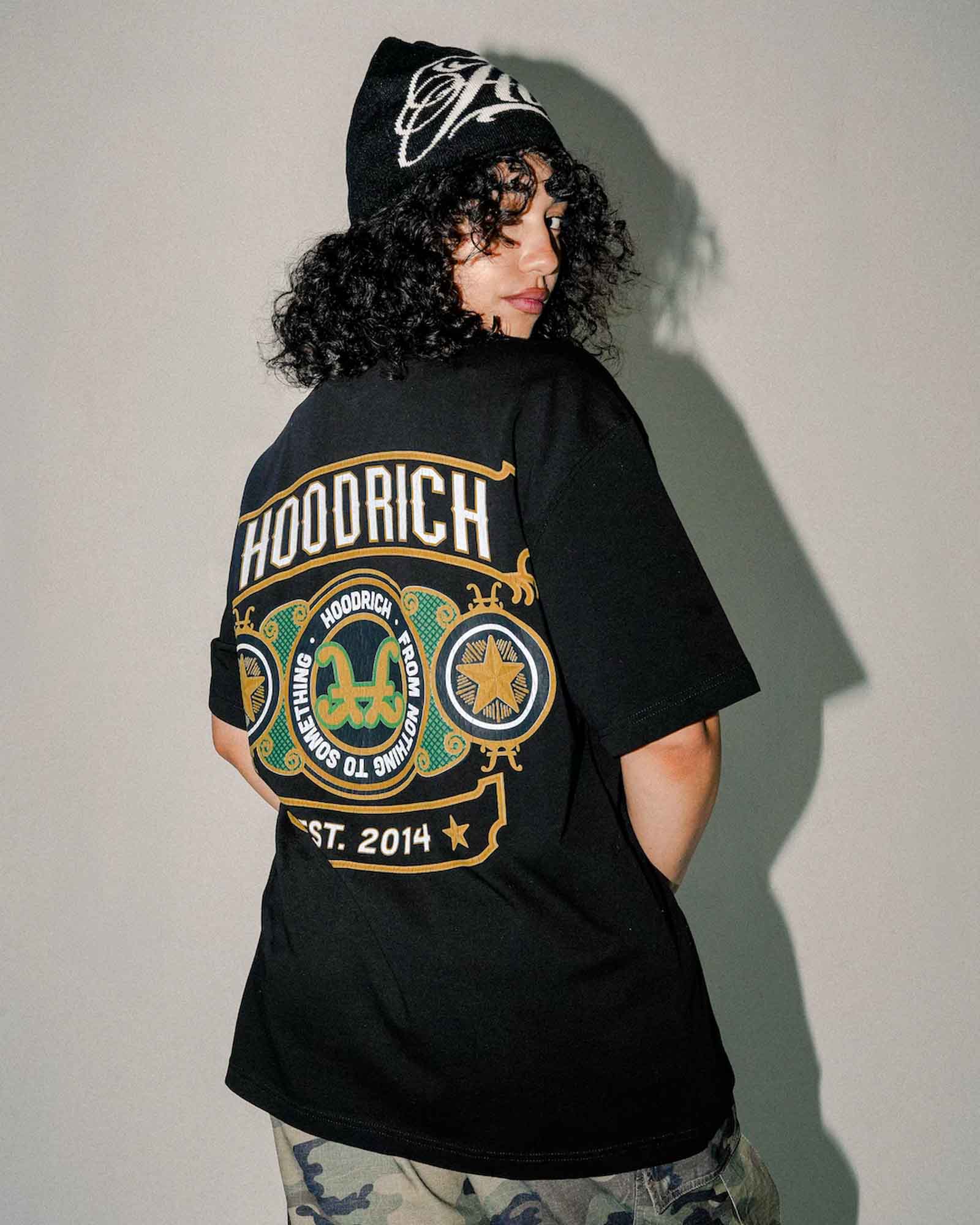 A person shown from the back wearing a black oversized HOODRICH t-shirt with a large multicolor graphic print, a black beanie, and camouflage pants, standing against a plain gray wall.