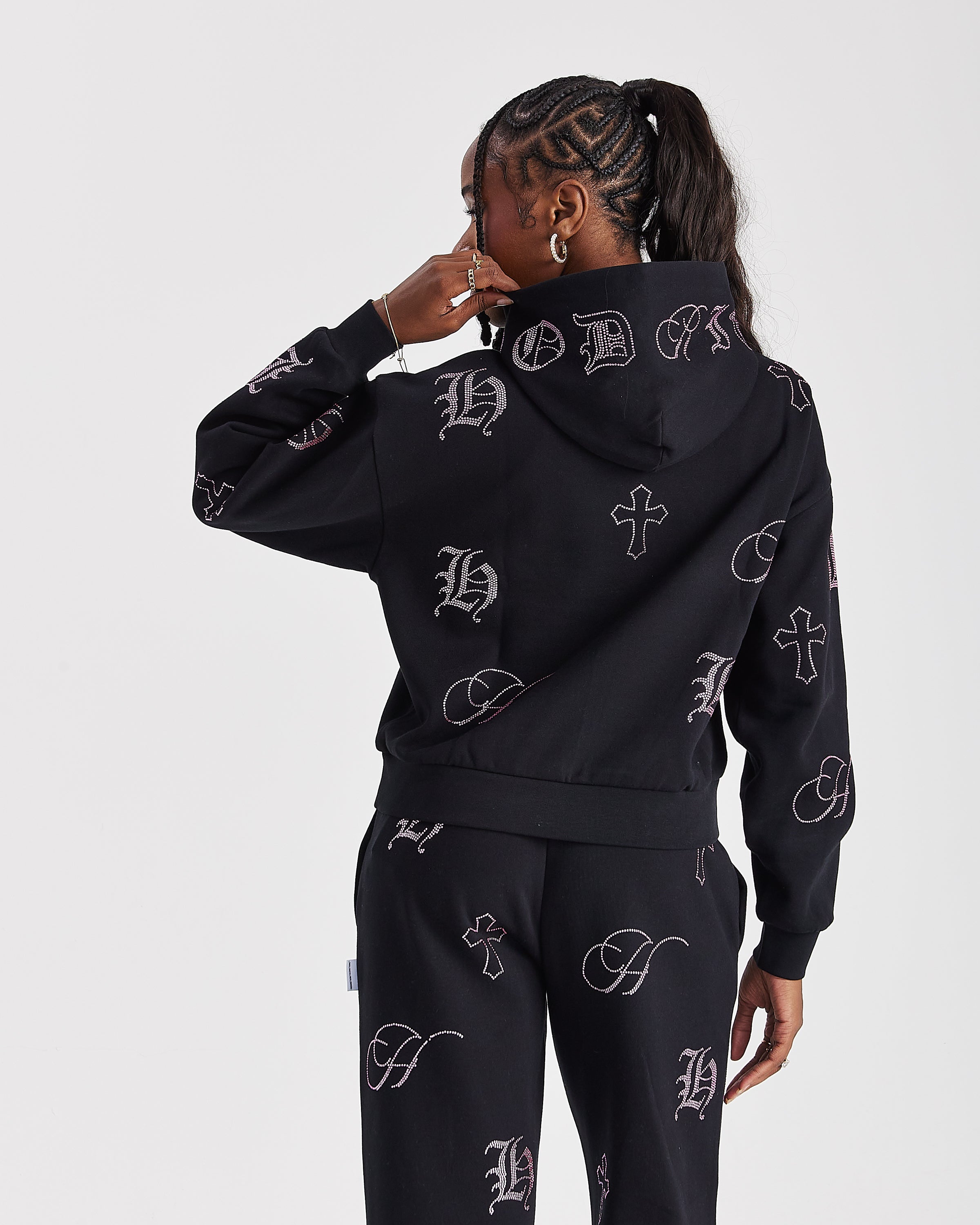 A model shown from the back wearing a black hooded sweatshirt and matching sweatpants set, both featuring an all-over light gray Gothic letter 'H' and cross pattern, against a light gray studio background.