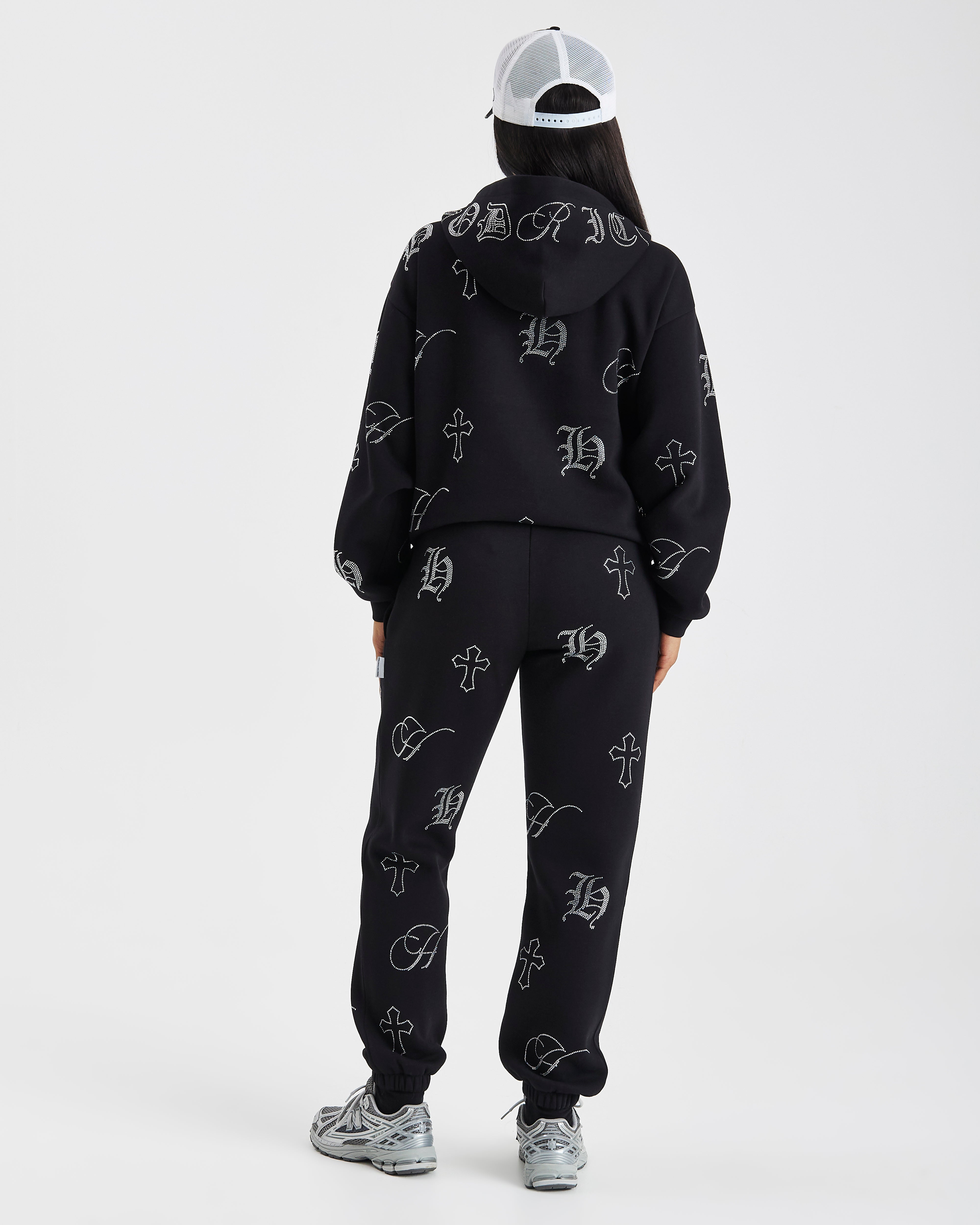 A model viewed from behind wearing a black hooded sweatshirt and matching jogger pants set, both featuring an all-over white gothic monogram pattern. The model is also wearing a white and black trucker cap and gray sneakers, set against a plain light gray studio background.