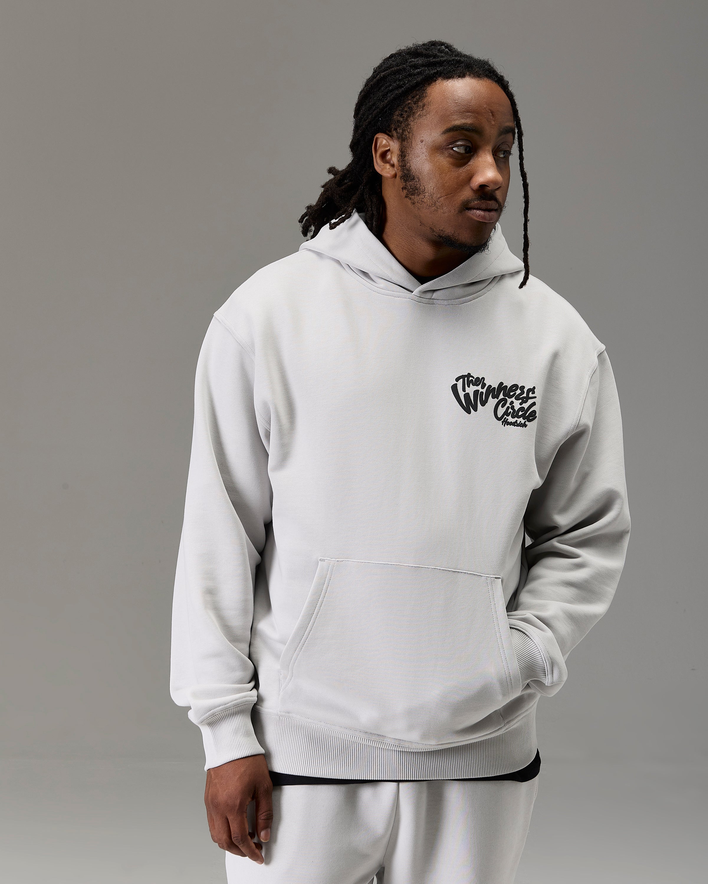 A man wearing a light gray pullover hoodie with a black 'The Winners Circle' graphic on the left chest, paired with matching light gray sweatpants, against a solid gray studio background.