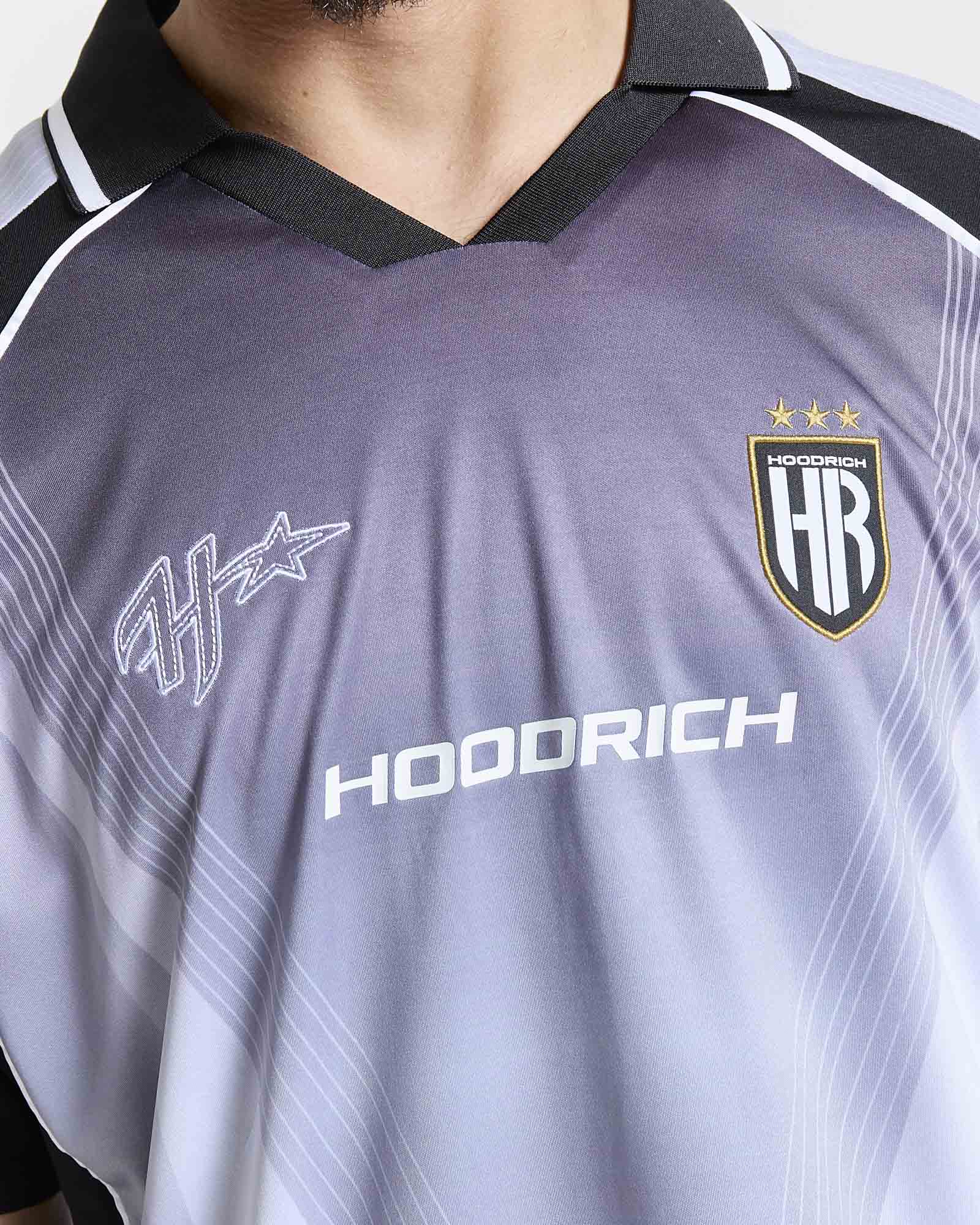 Close-up of a HOODRICH soccer jersey with a dark gray and light blue gradient, featuring the HOODRICH sponsor logo, the team crest, and a stylized star logo on the chest.